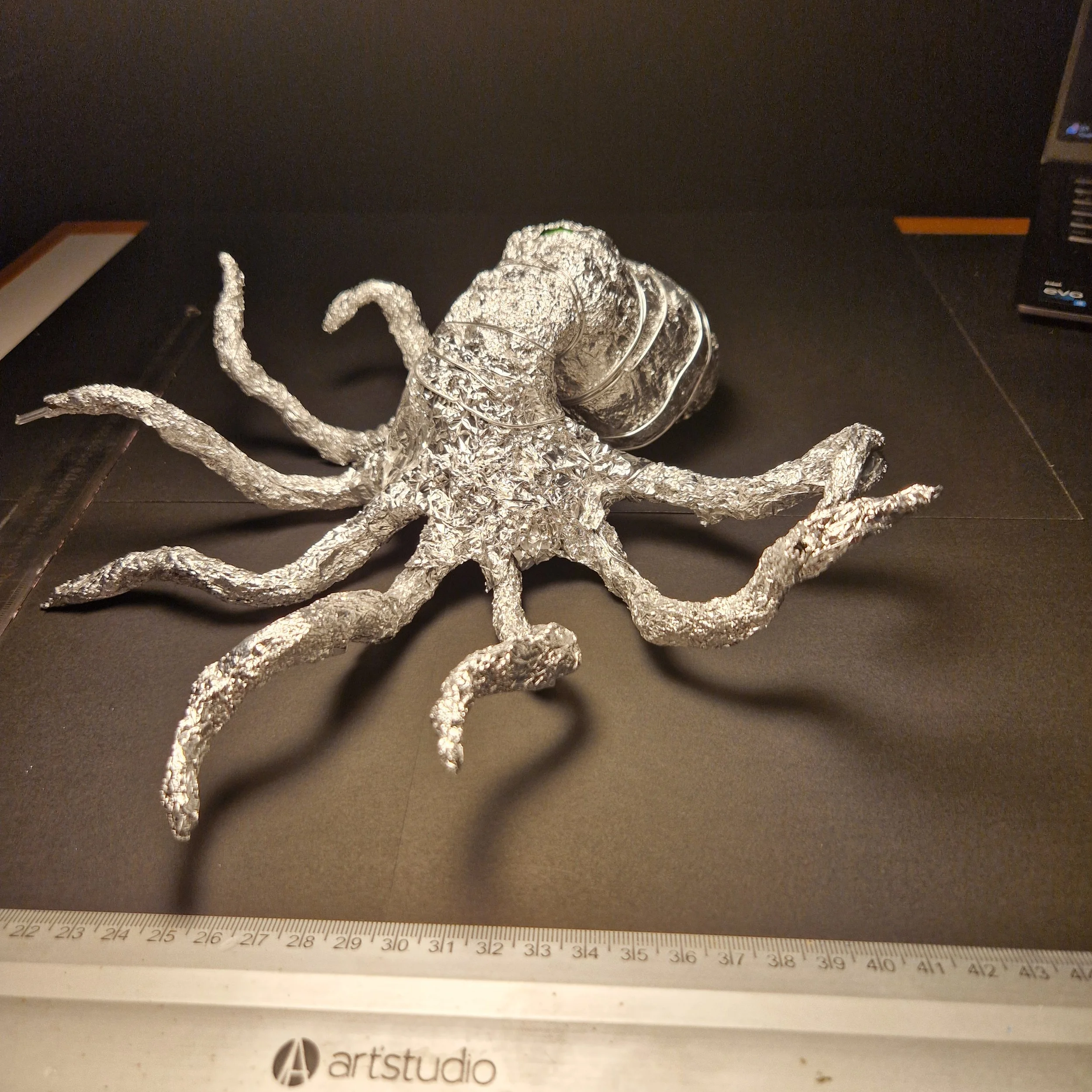 A crumpled aluminum foil sculpture resembling a fantastical creature with multiple tentacle-like appendages, a curled tail, and other intricate details, placed on a black surface with a ruler and a partial box nearby.