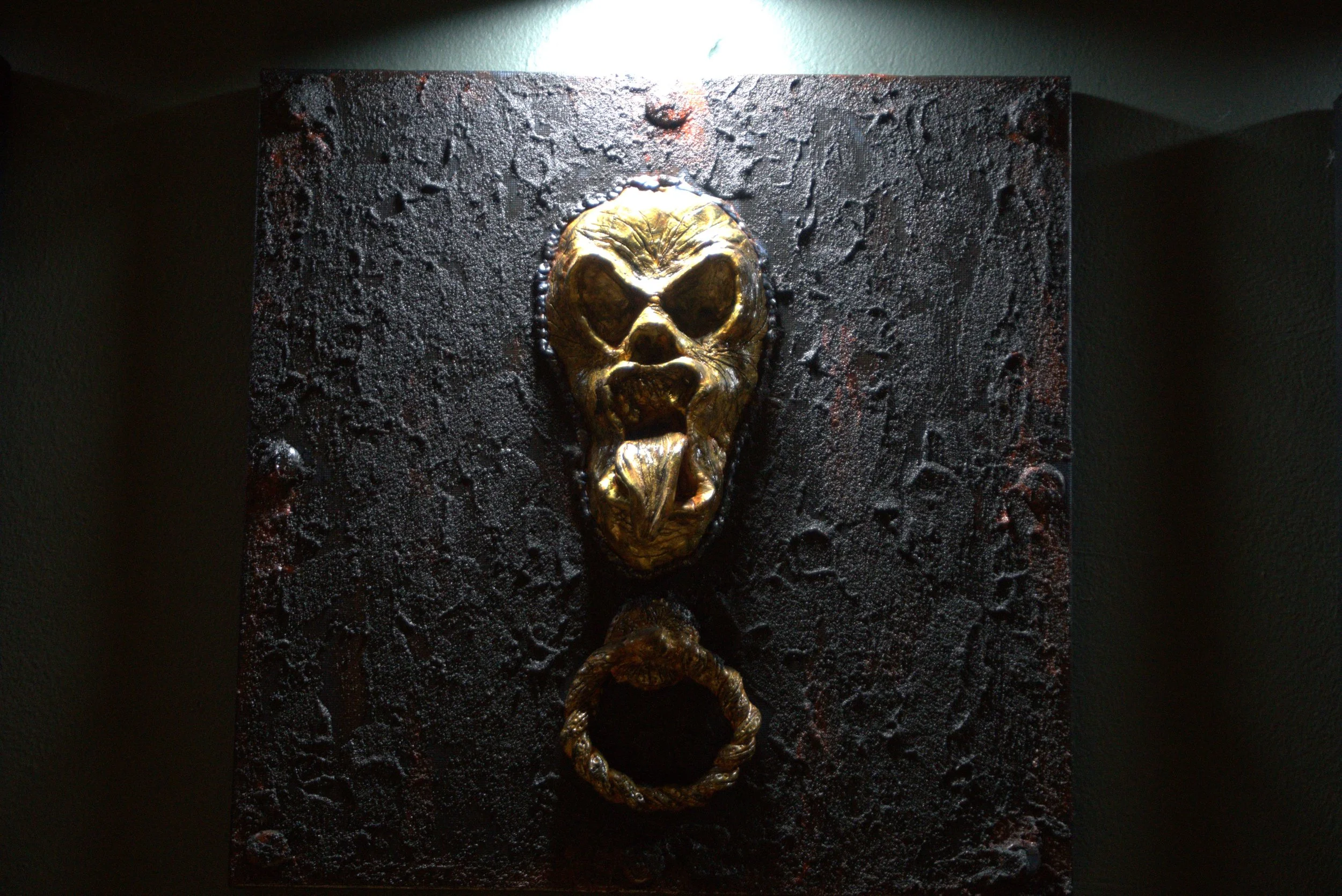 A textured black wall with a central metallic golden skull ornament and a looped metallic chain hanging below it.