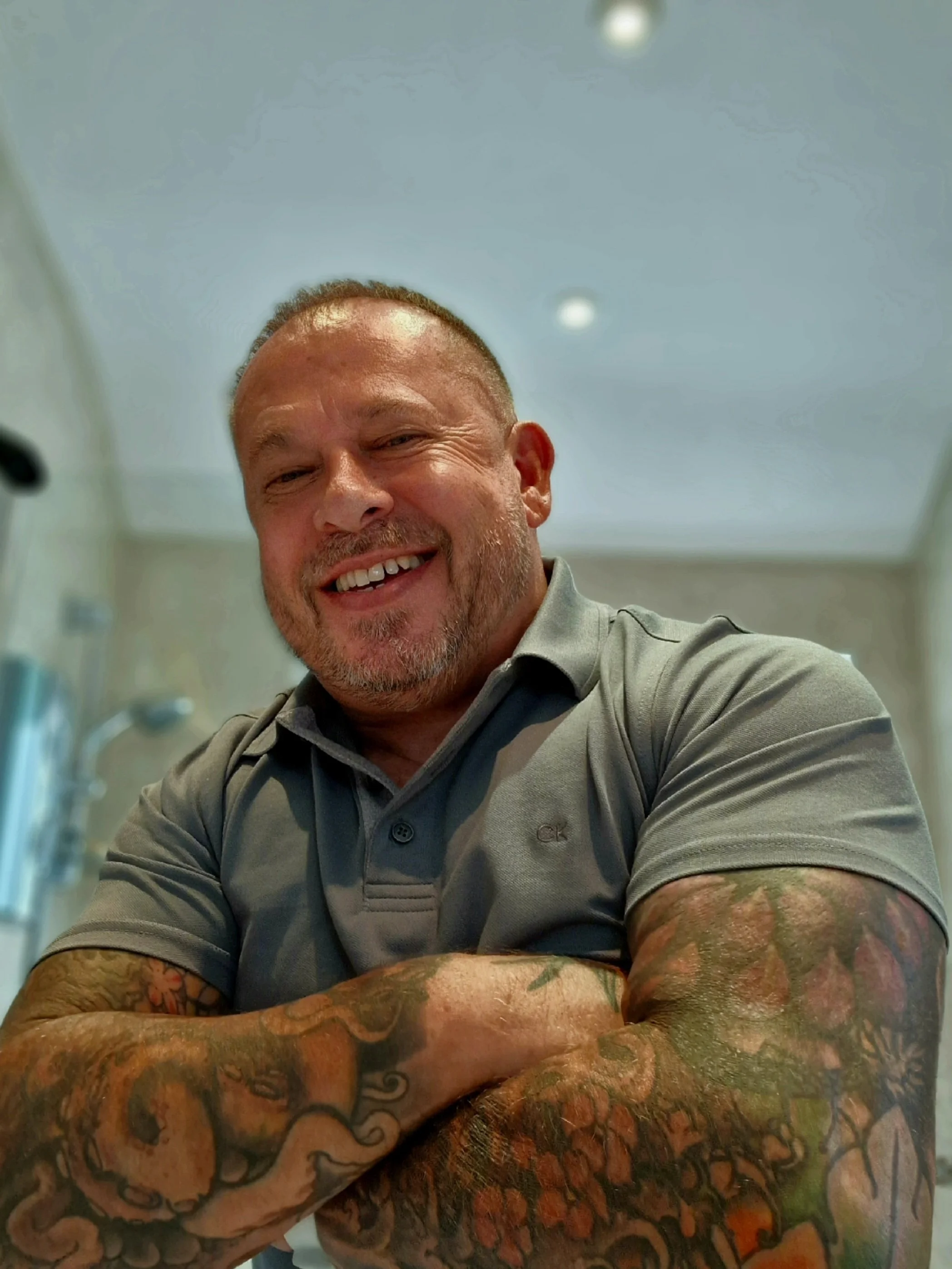 A smiling man with a shaved head and tattoos on his arms, wearing a gray polo shirt, sitting in a medical or professional setting, with a blurred background and ceiling lights.