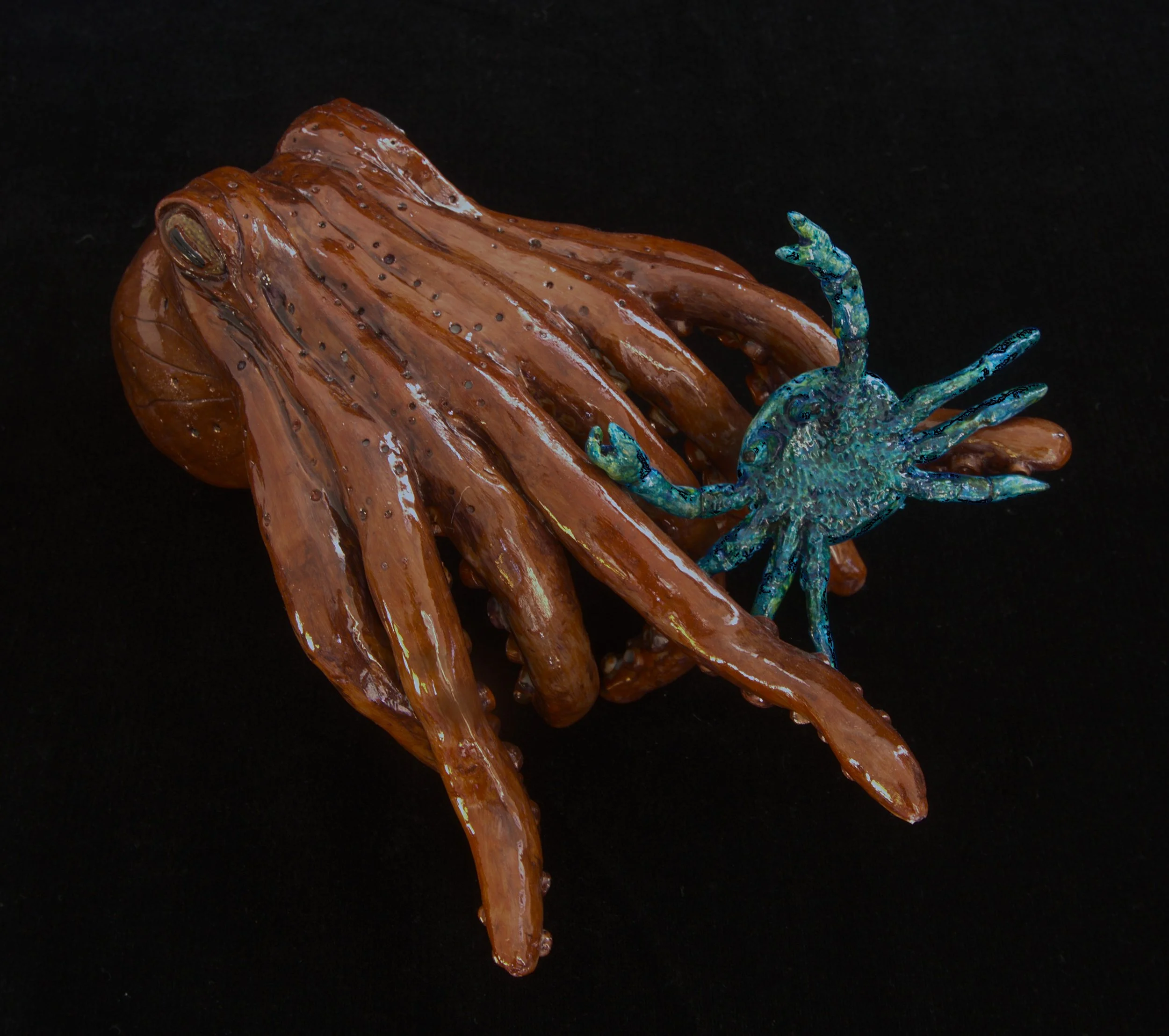 A decorative glass sculpture of a crab with a blue-green body and large claws, positioned on a polished, brown, wood-like structure with tiny black dots, against a solid black background.