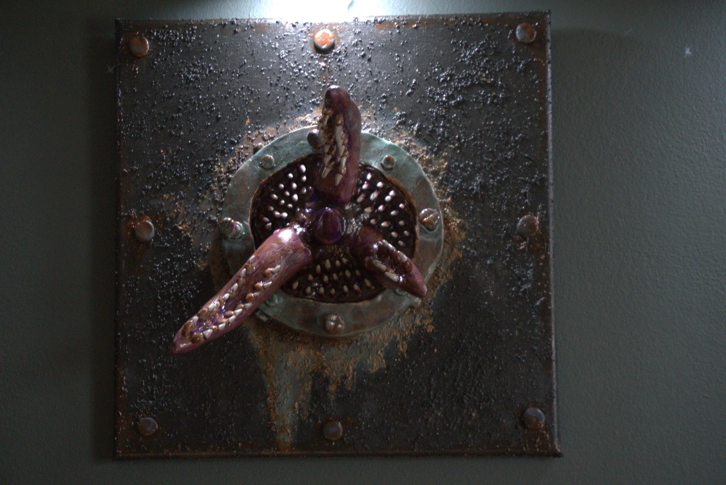 A handmade octopus sculpture mounted on a metal plate, attached to a dark, textured surface with screws.