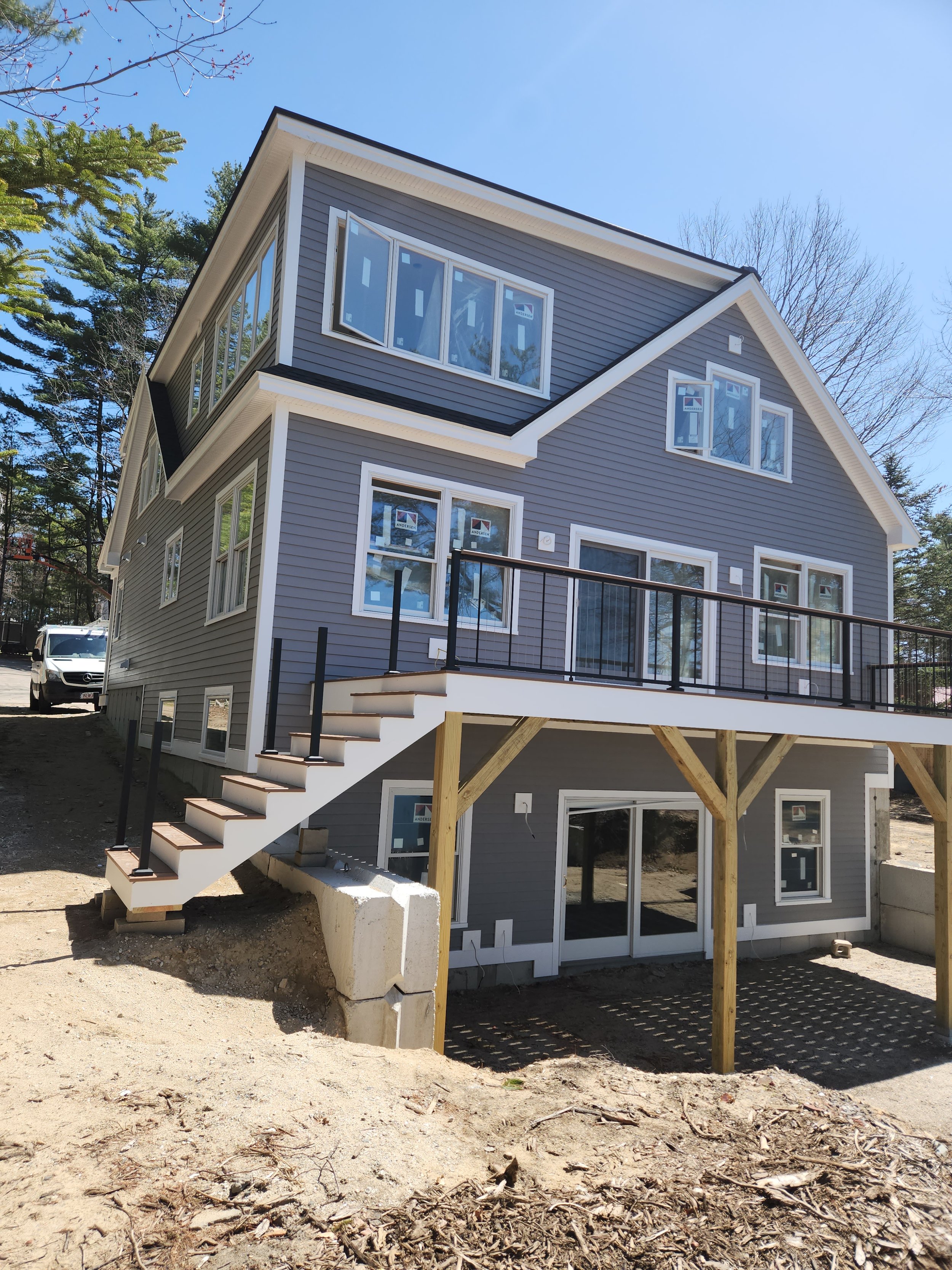 A house under construction with gray siding, multiple windows, a staircase leading to a upper-level deck, and support beams visible underneath.