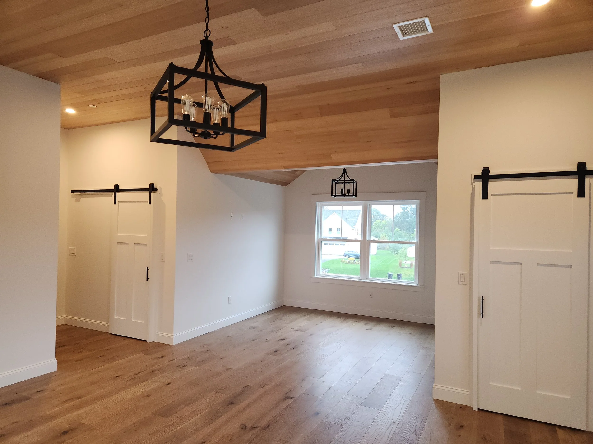 An interior living room area with wooden floors and ceiling, white walls, large window showing a view of suburb neighborhood, black metal chandelier, and sliding barn doors.