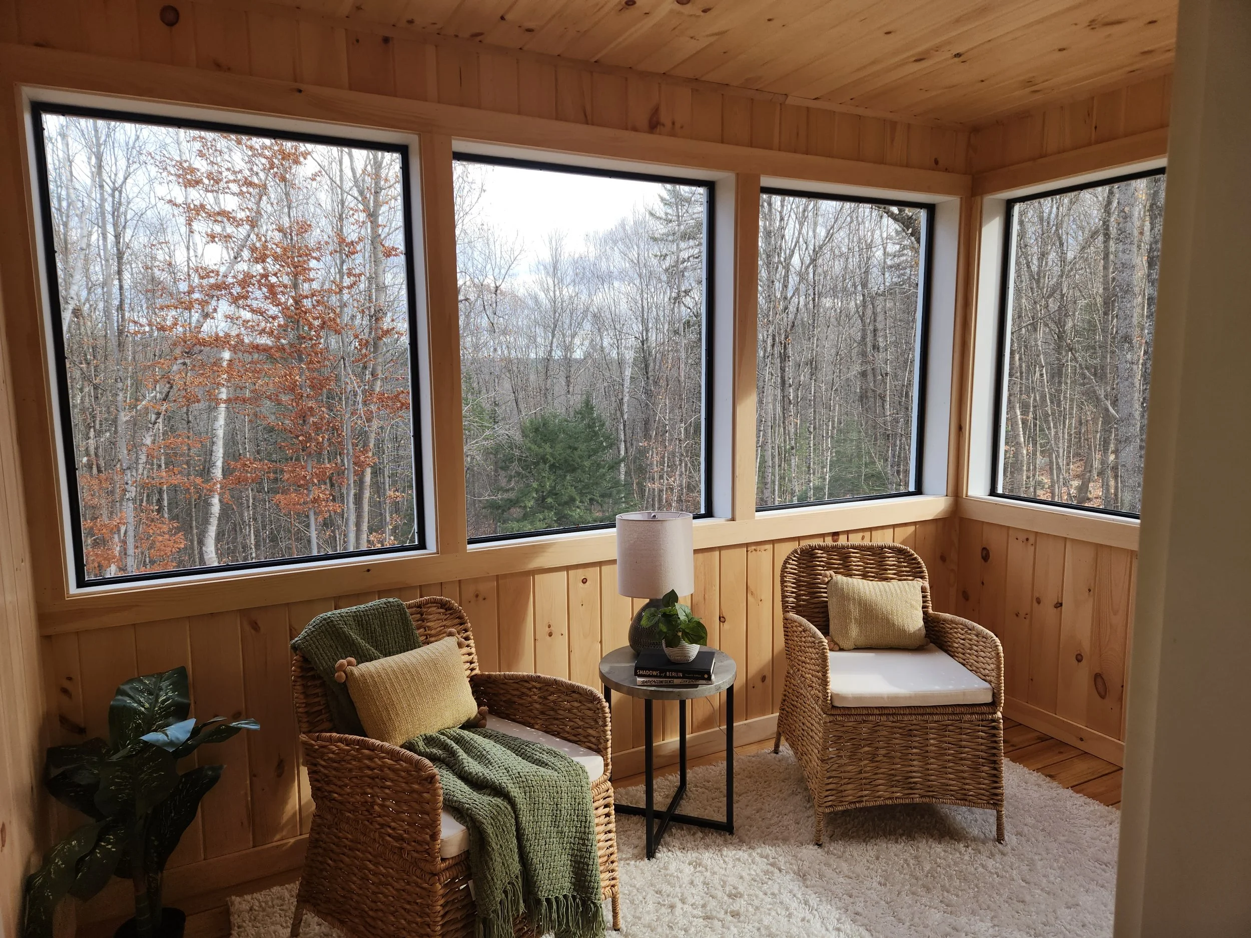 A cozy sunroom with wooden walls and large windows showing a forested landscape outside. There are four wicker chairs with cushions, a small round table with books, a lamp, and a plant, all set on a plush beige rug.