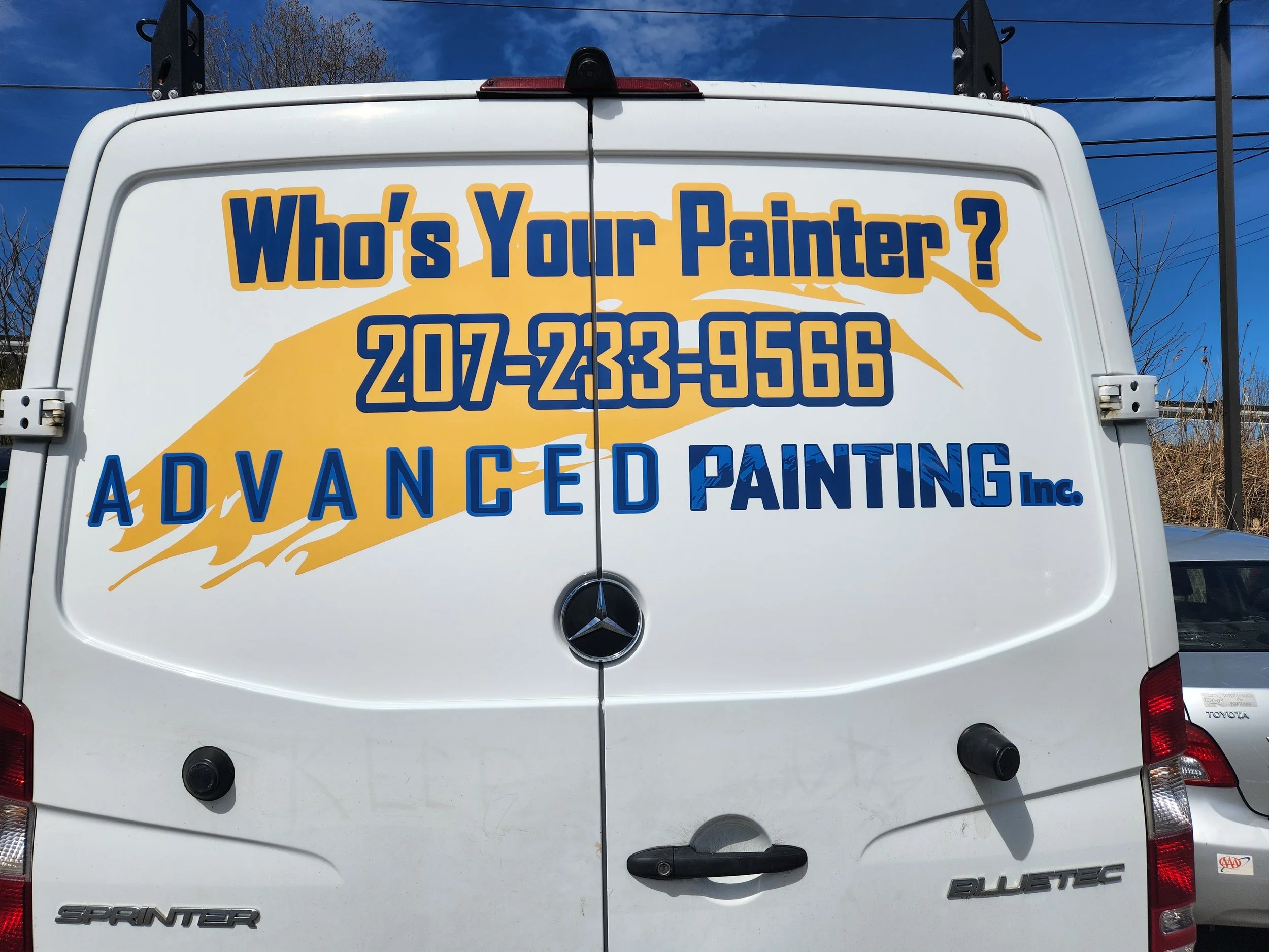 The back of a white van with yellow and blue text advertising a painting service. The text reads: "What's Your Painter?" along with a phone number and company name, "Advanced Painting Inc." The van has a Mercedes-Benz logo and is parked outdoors under a clear blue sky.