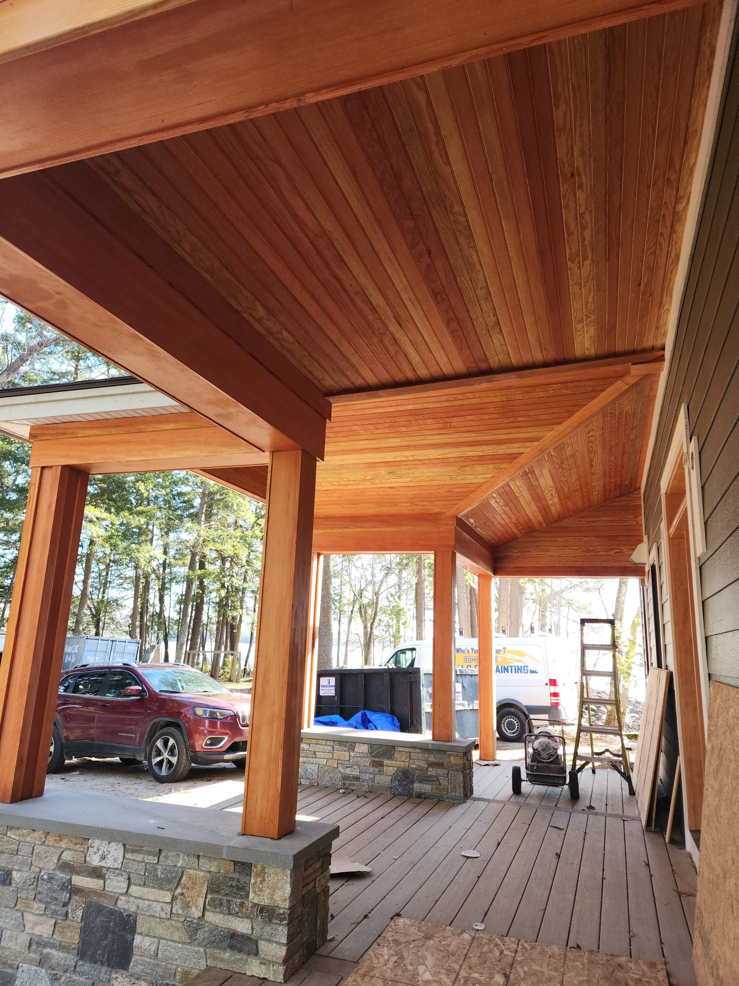 Wooden porch with support beams, a stone and brick column at the base, and construction tools and vehicles in the background.