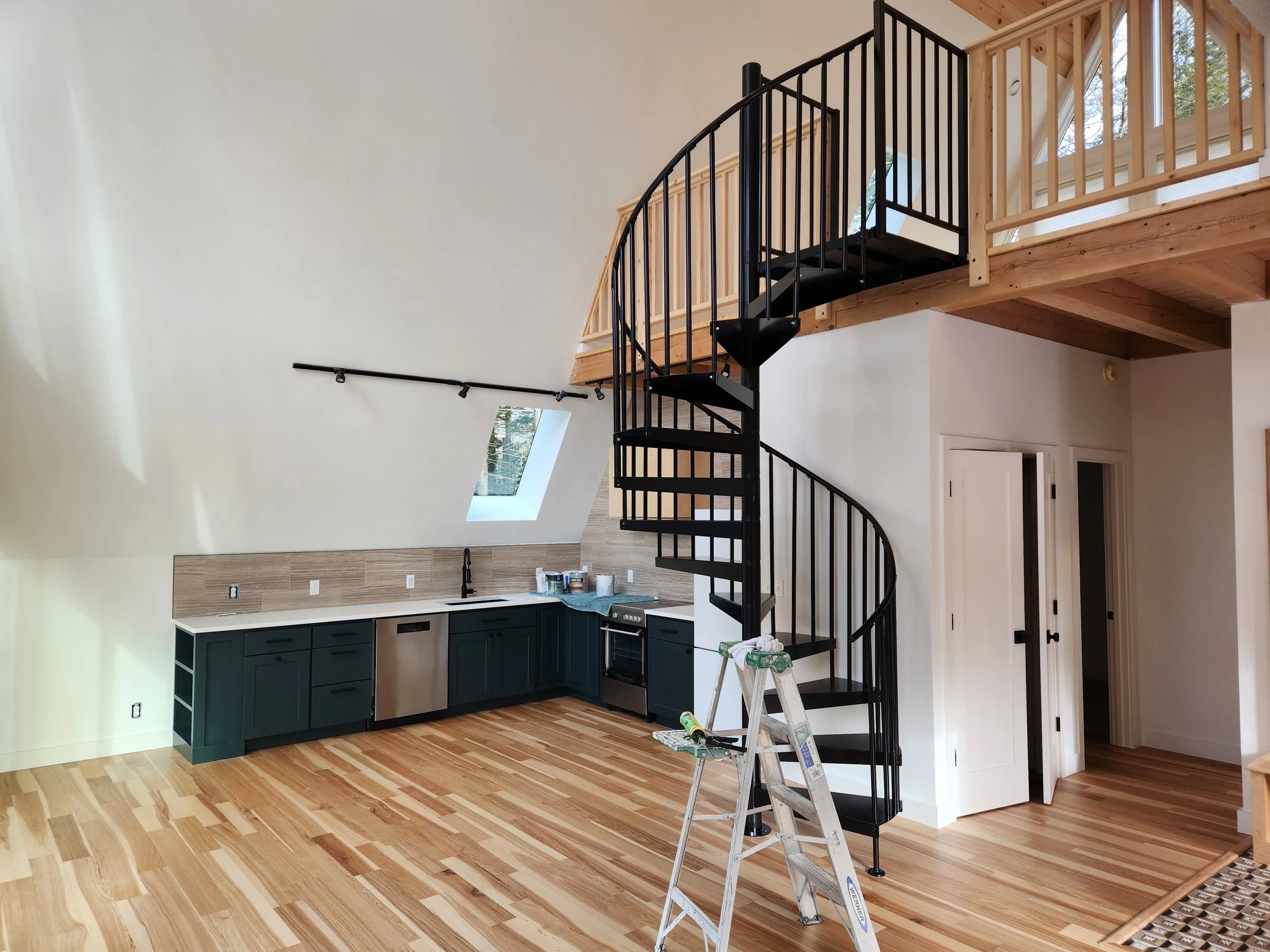 Interior of a house under construction featuring a spiral staircase with black railings and a staircase with wooden railings, kitchen cabinets, and hardwood flooring.