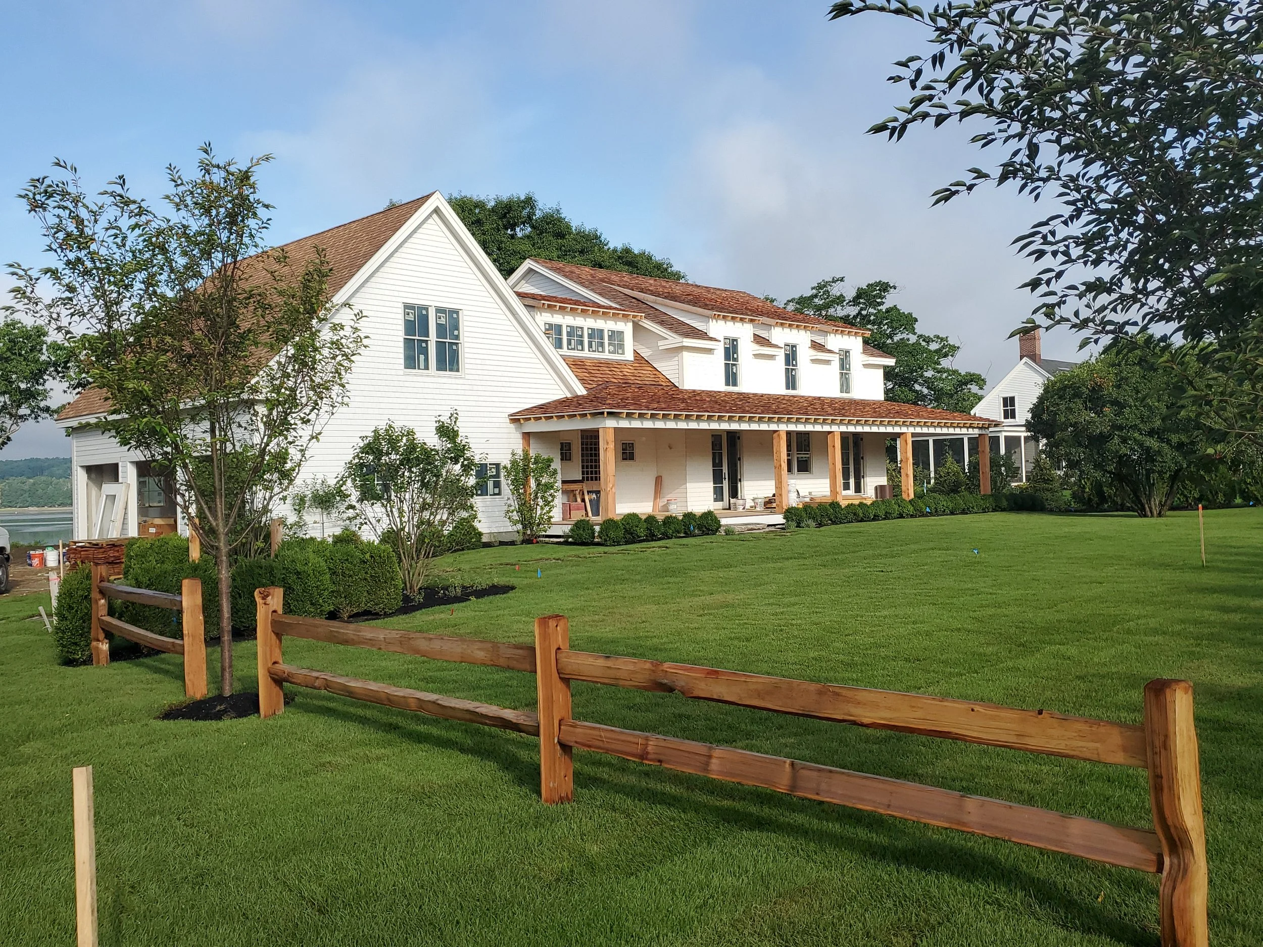 A white house with a brown roof under construction, surrounded by green trees and a well-maintained lawn, with a wooden fence in the foreground.
