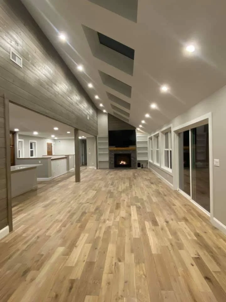 Spacious living room with hardwood floors, a stone fireplace, large windows, and a sloped ceiling with recessed lighting.