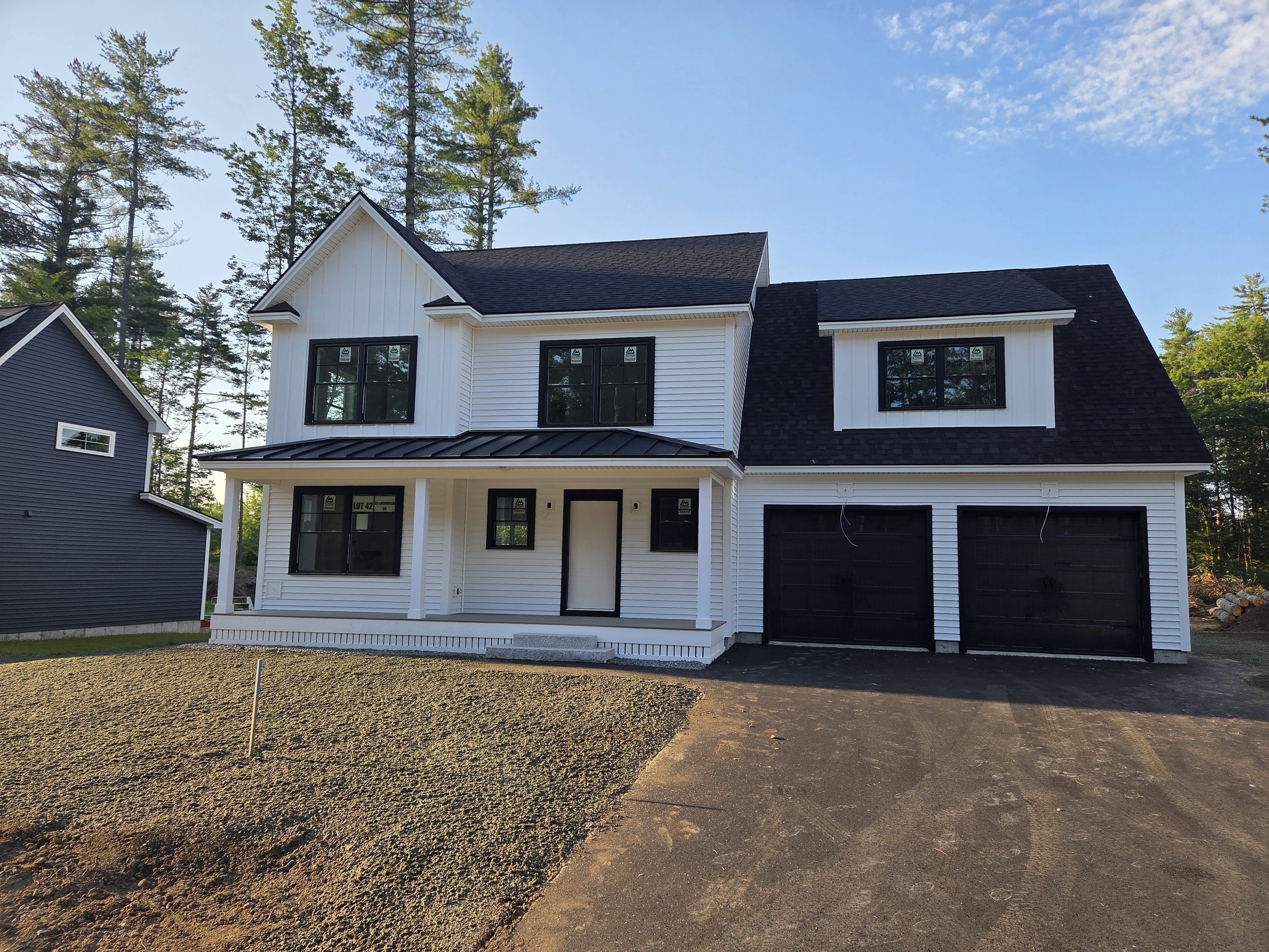 A partially constructed white two-story house with black framed windows and a black roof, surrounded by tall trees and a dirt yard.