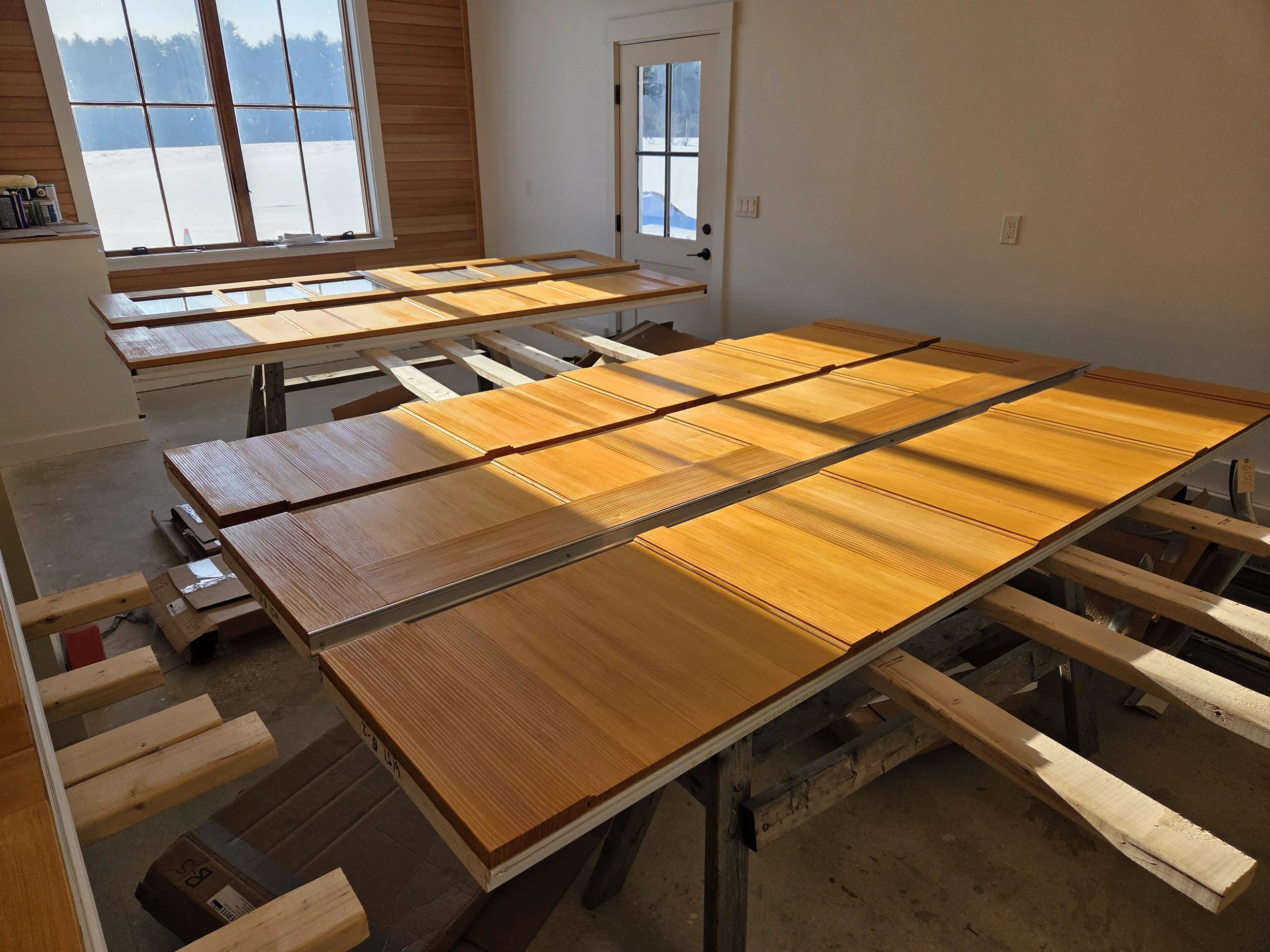 Interior of a room with large windows showing snow outside. Several wooden cabinet doors are laid face down on tables, likely for finishing or assembly. The room appears to be under construction or renovation.