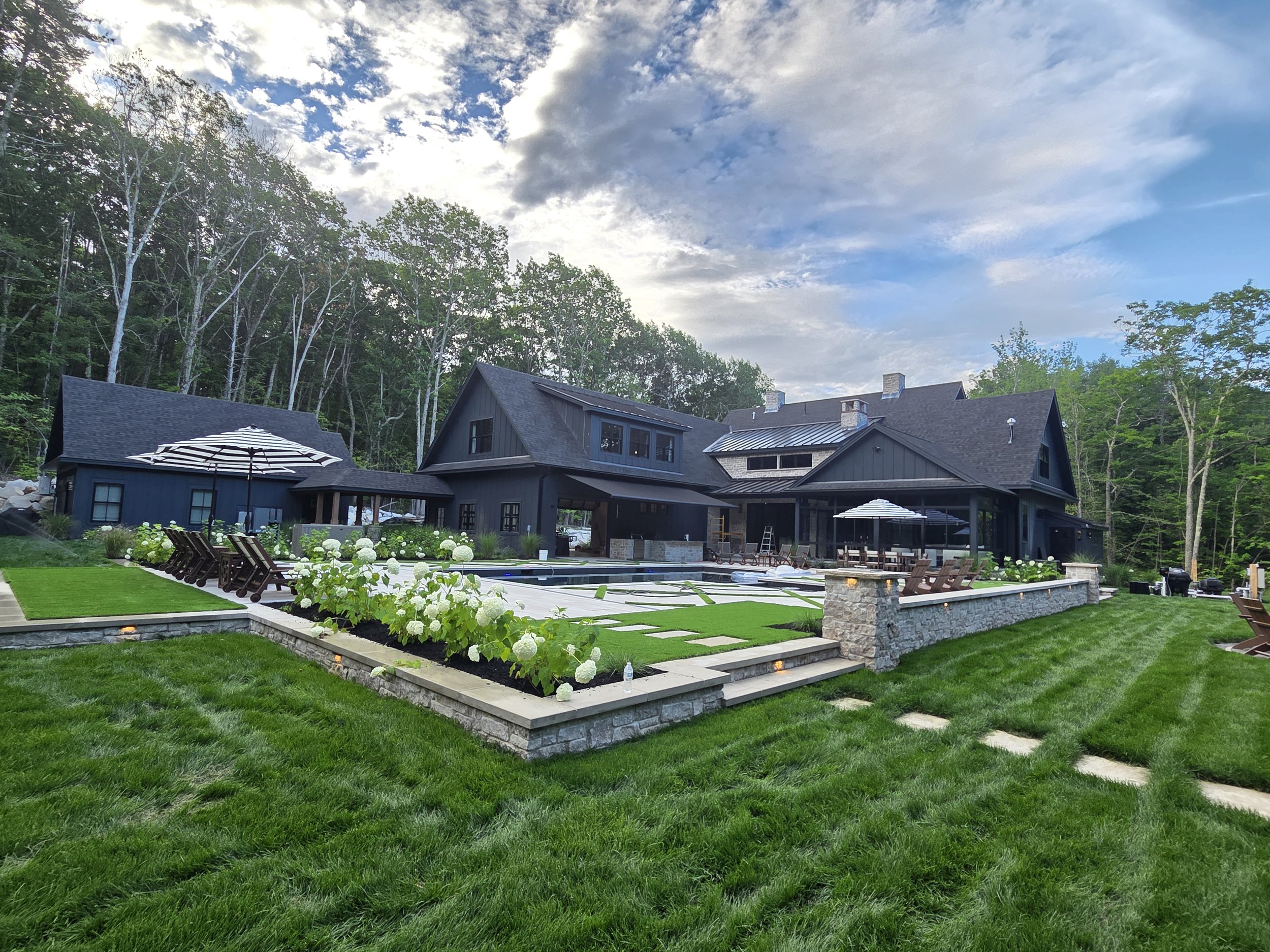 A modern house with dark exterior, large windows, and a metal roof, surrounded by lush green lawn and landscaping, with a cloudy sky overhead.