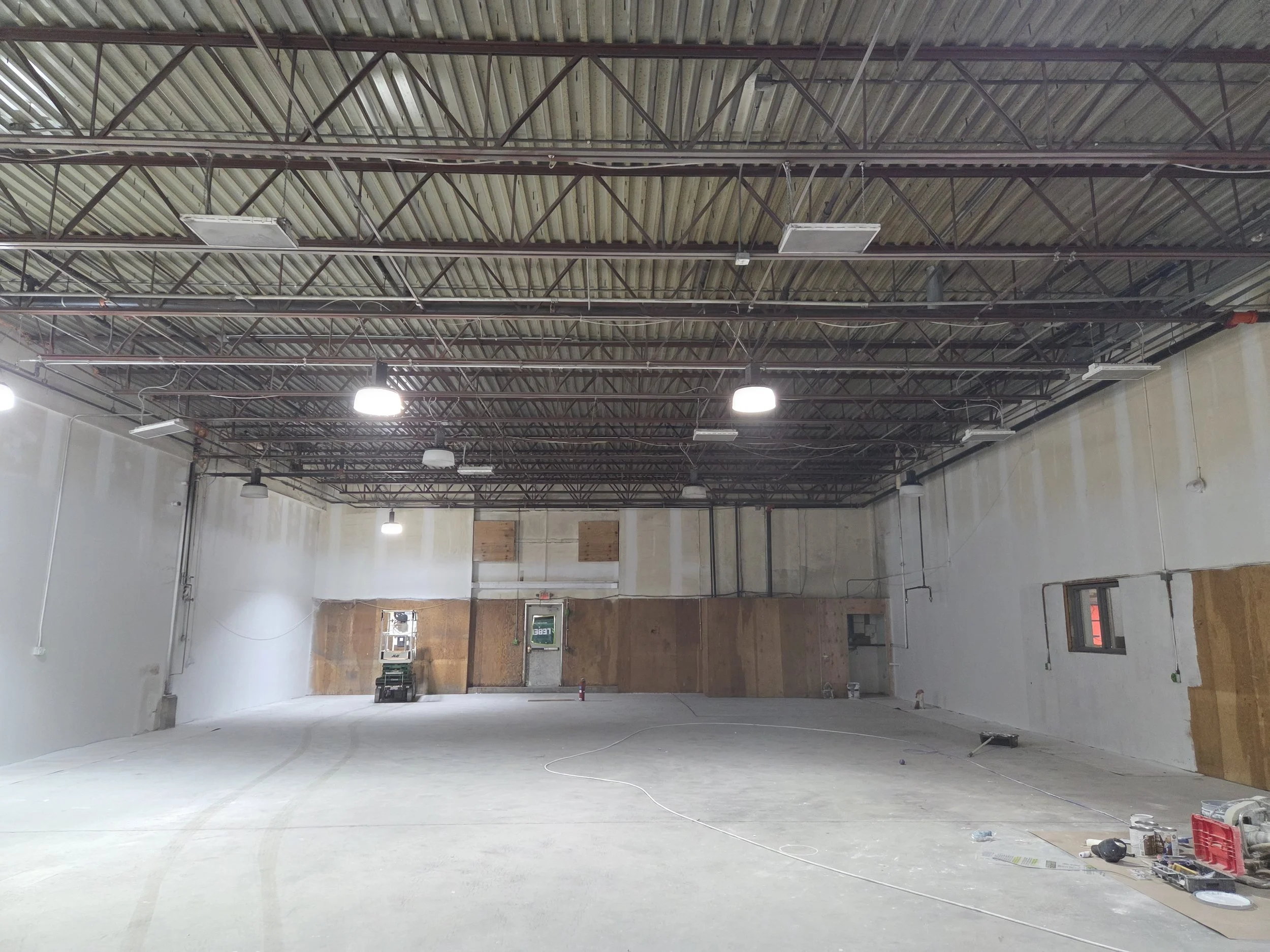 Interior of a large, unfinished commercial or industrial building with a high metal roof, exposed trusses, suspended lighting fixtures, and unfinished walls with some construction tools and materials on the floor.