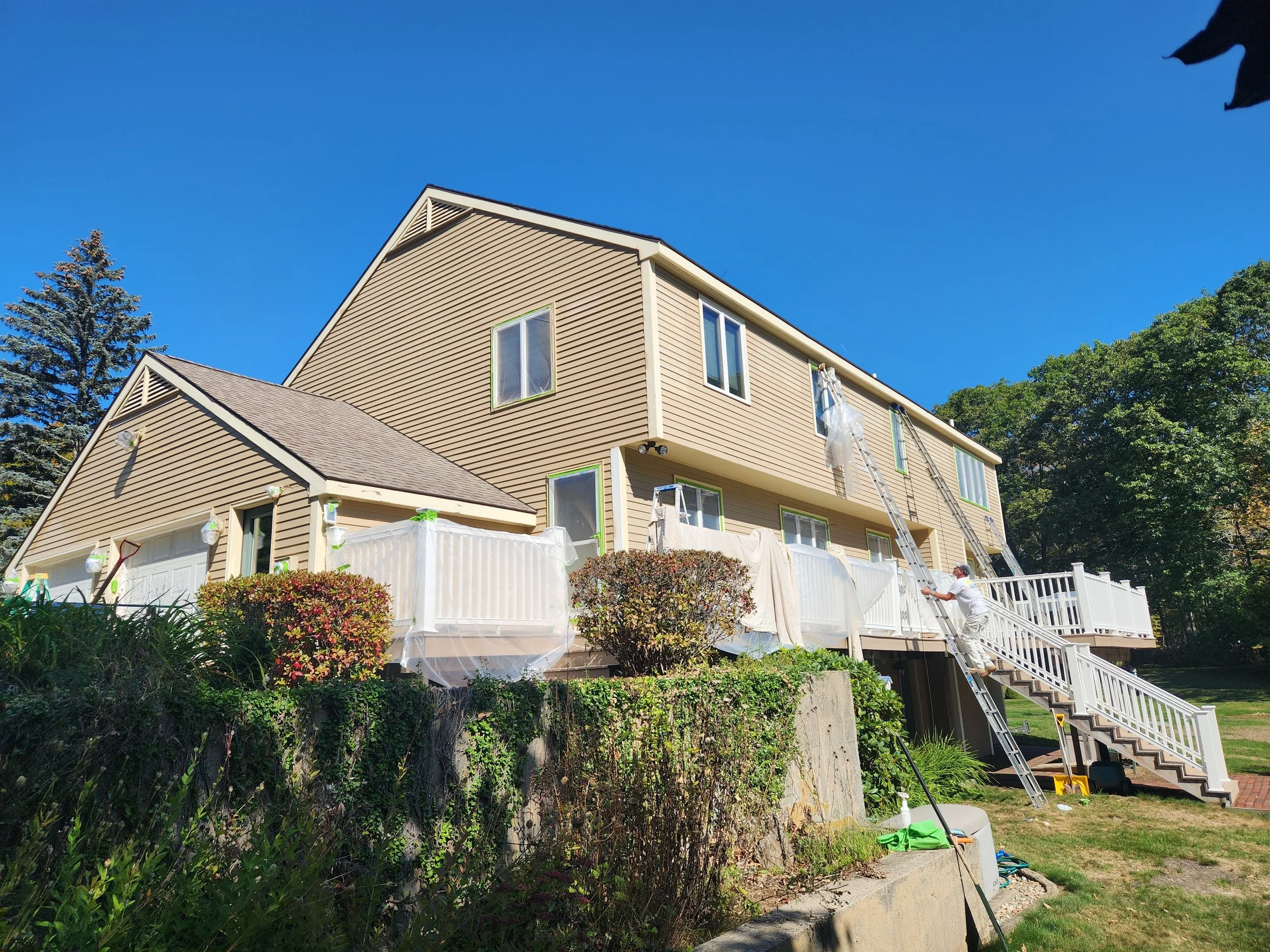 A multi-story house with beige siding, multiple windows, and a white staircase outside. Two ladders are leaning against the house, and two workers are near the bottom of the stairs, possibly performing maintenance or renovation. There is a fence, bus
