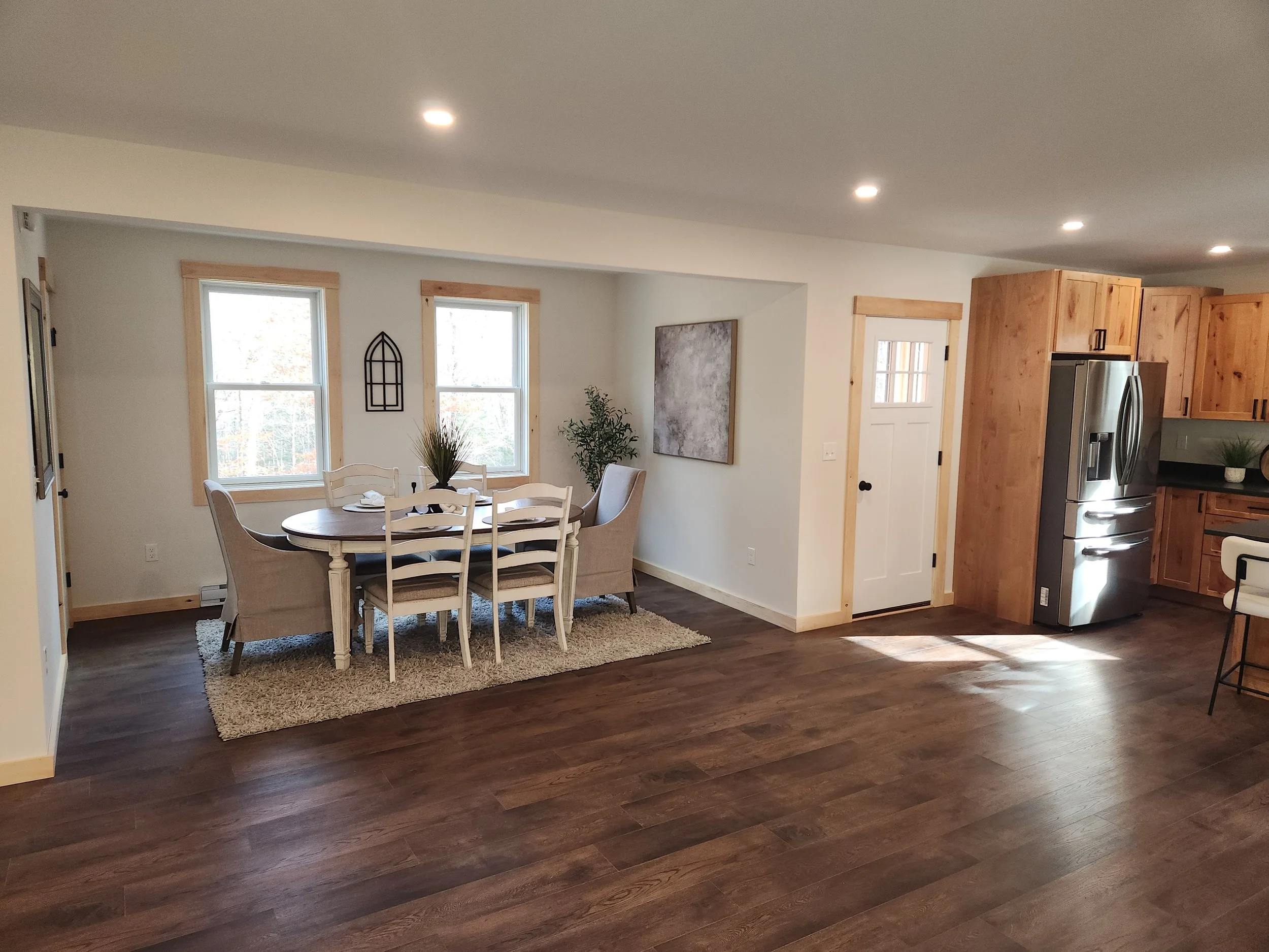 Interior of a modern house with hardwood floors, a dining table with chairs, large windows, a wall hanging, and kitchen cabinets with a refrigerator.