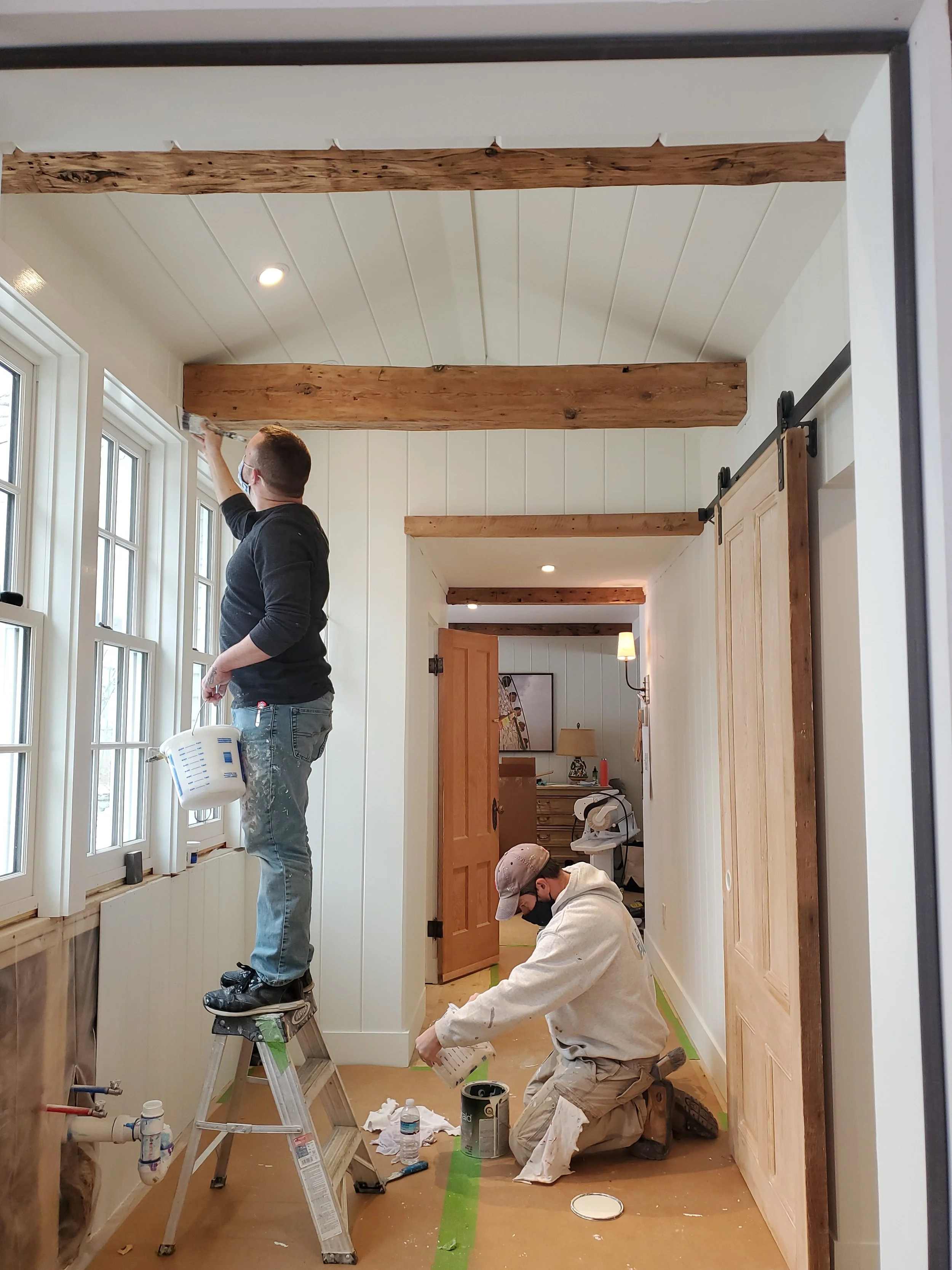 Two people painting the walls of a room with white beadboard paneling, natural wood beams on the ceiling, and wood accents on the doors.