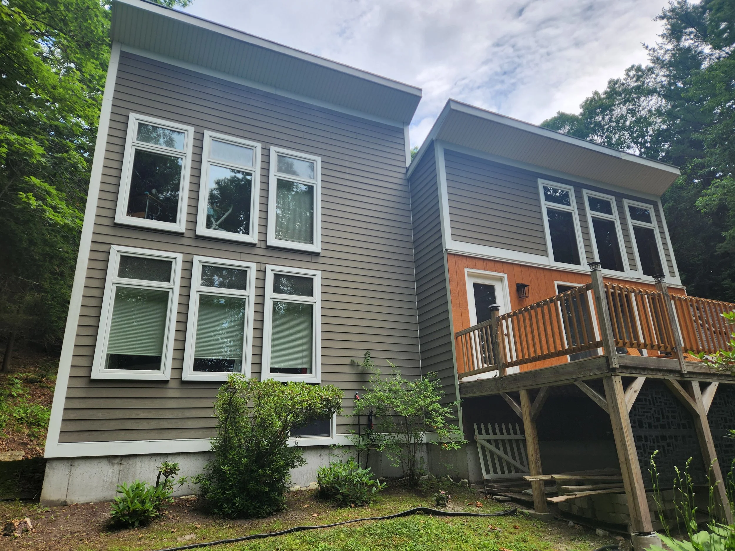 The back of a modern multi-story house with gray siding and white trim windows, a deck with a wooden railing, and a garden with small bushes, with a cloudy sky and trees in the background.