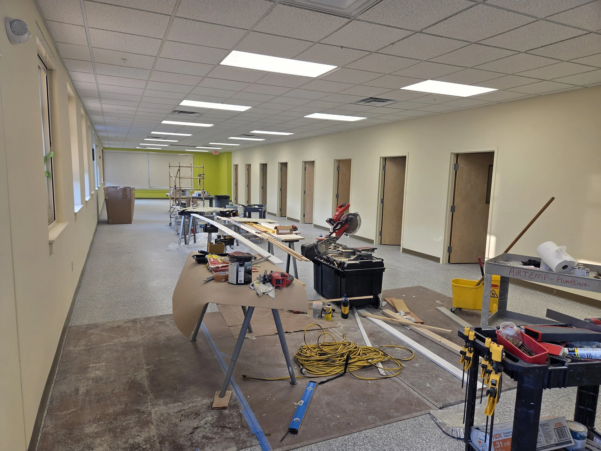 Interior of a building under construction or renovation with tools, scaffolding, and construction materials such as paint cans, power tools, ladders, and extension cords scattered around. Multiple room doorways are visible on the right side, and a la
