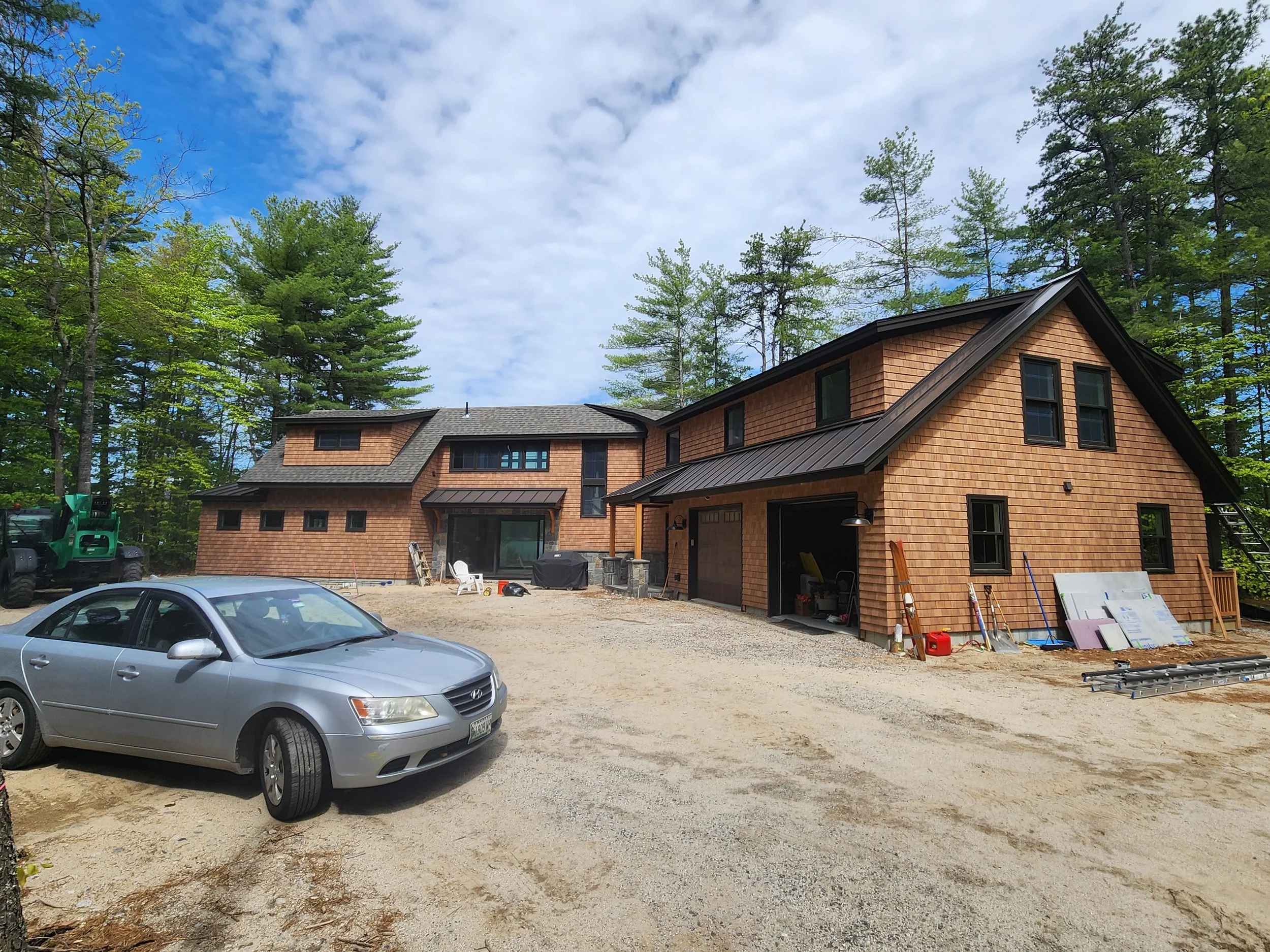 Newly constructed house with wooden shingles, surrounded by green trees, under a partly cloudy sky. Construction materials and a silver car parked in the front yard, with construction tools and equipment nearby.