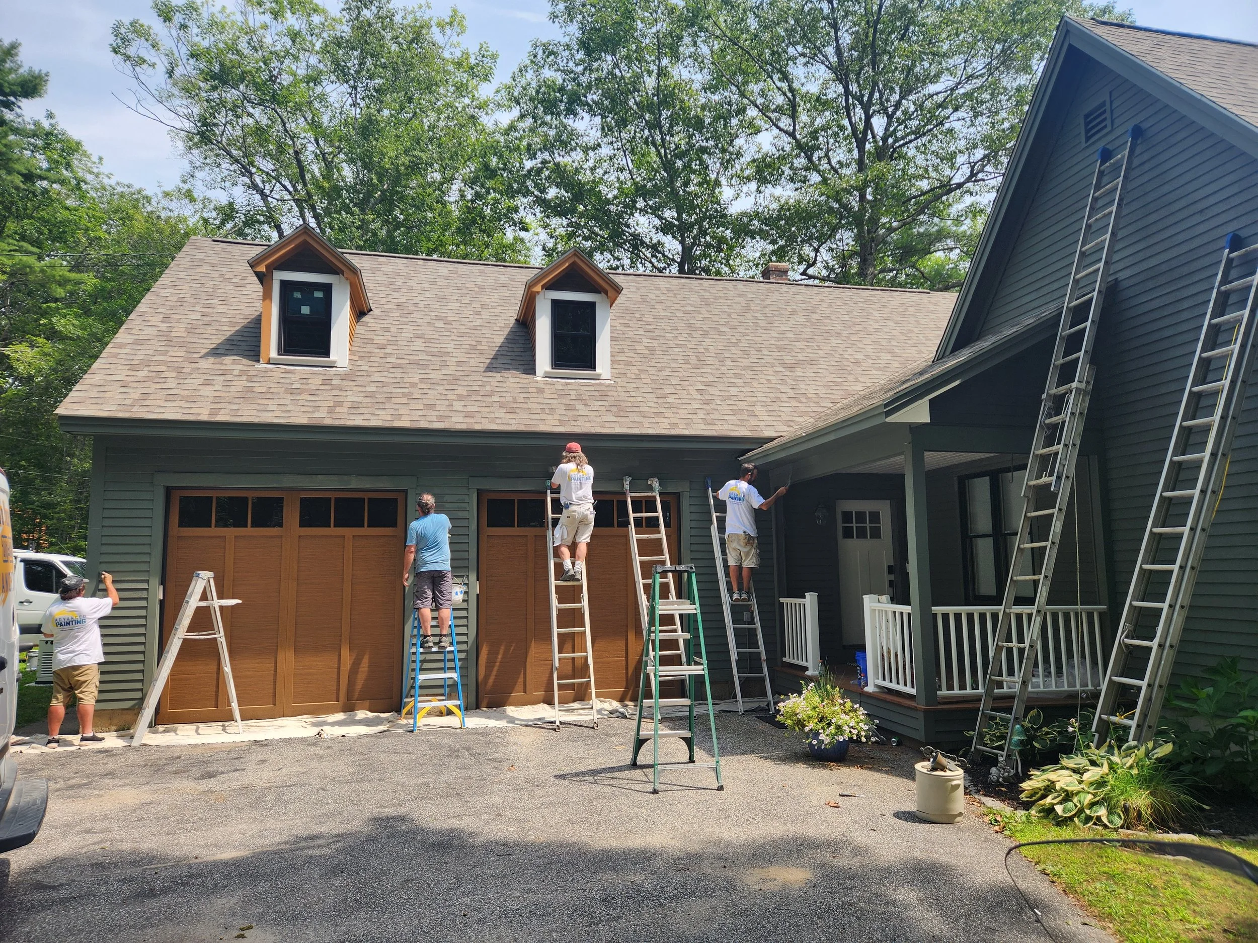 A home construction scene with several workers painting the house exterior. The house has a tan-colored garage door, siding, and two dormer windows on the roof. Ladders, scaffolding, and painting equipment are visible, and the workers are wearing saf