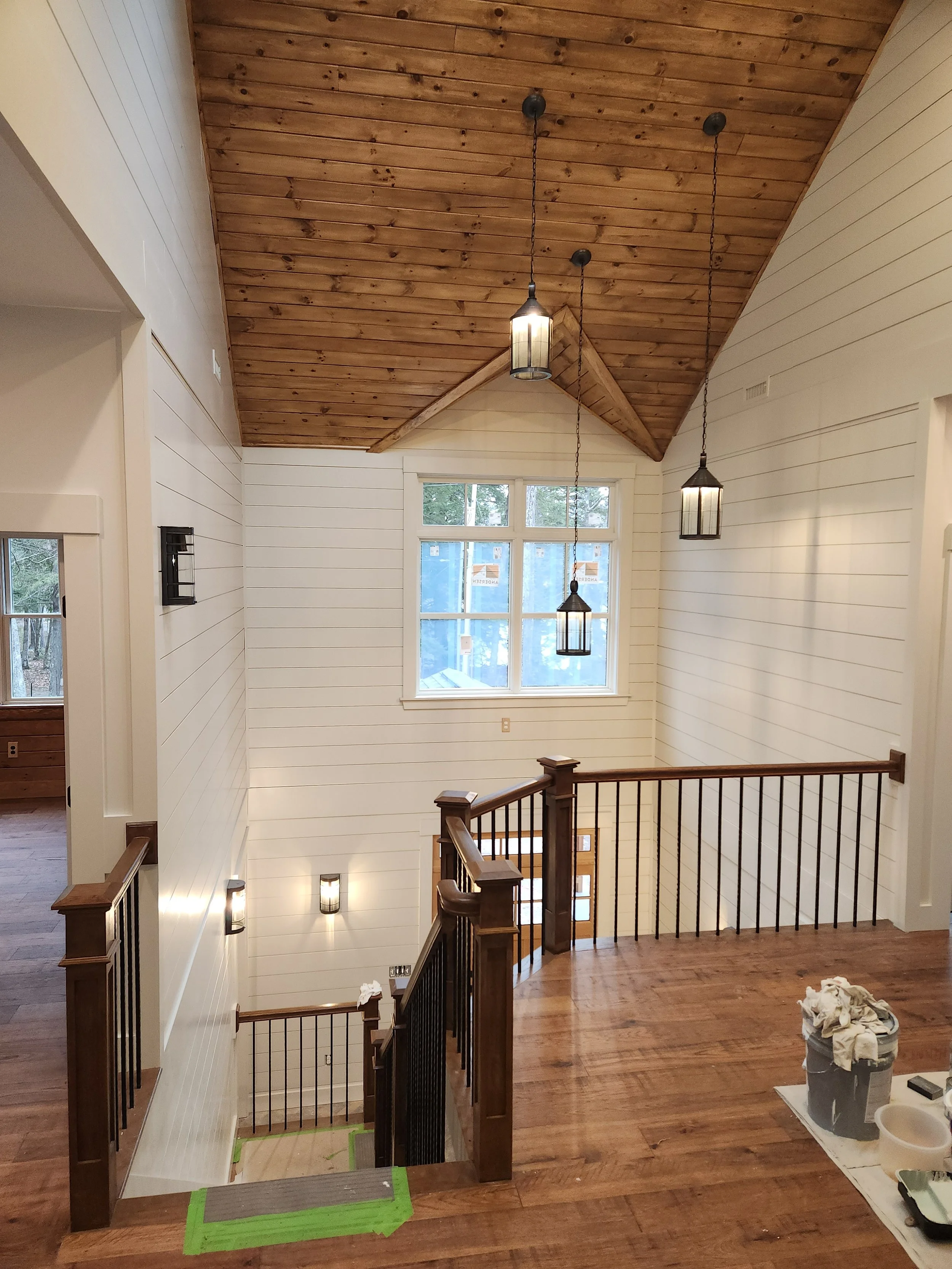 Interior view of a staircase landing with white shiplap walls, a large window, and a wooden ceiling with pendant lights. There are some construction items on a cloth on the floor to the right.
