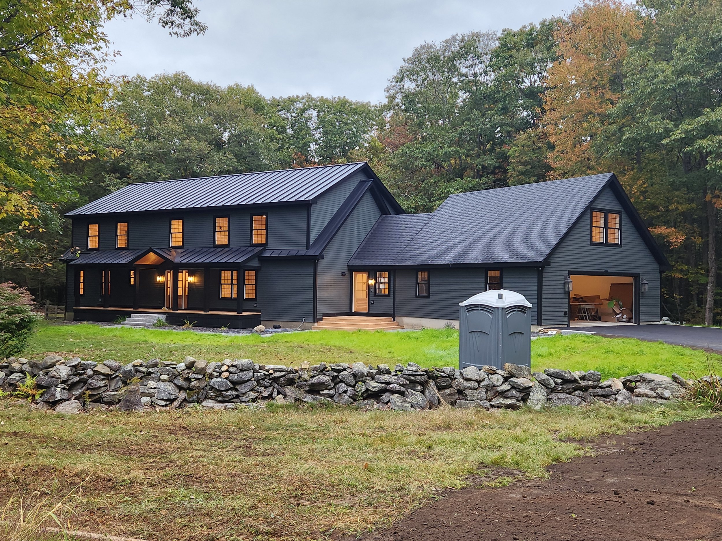 A modern two-story house with dark exterior siding, multiple windows glowing with warm light, a front porch with stairs, surrounded by trees with fall foliage, and a neatly maintained lawn and stone wall in the foreground.