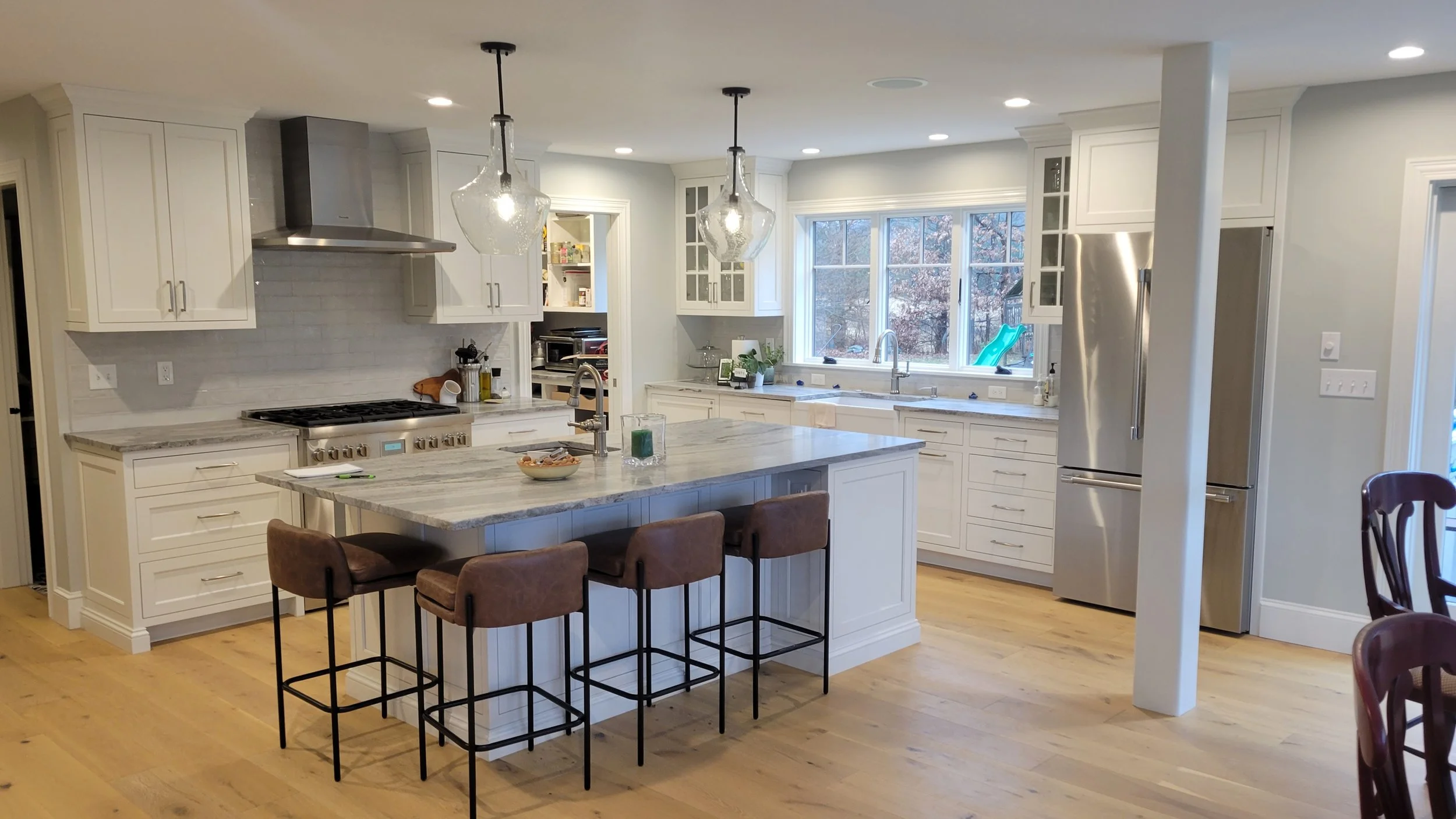 White kitchen with island, brown chairs, stainless steel refrigerator, large window, pendant lights, and light wood flooring.