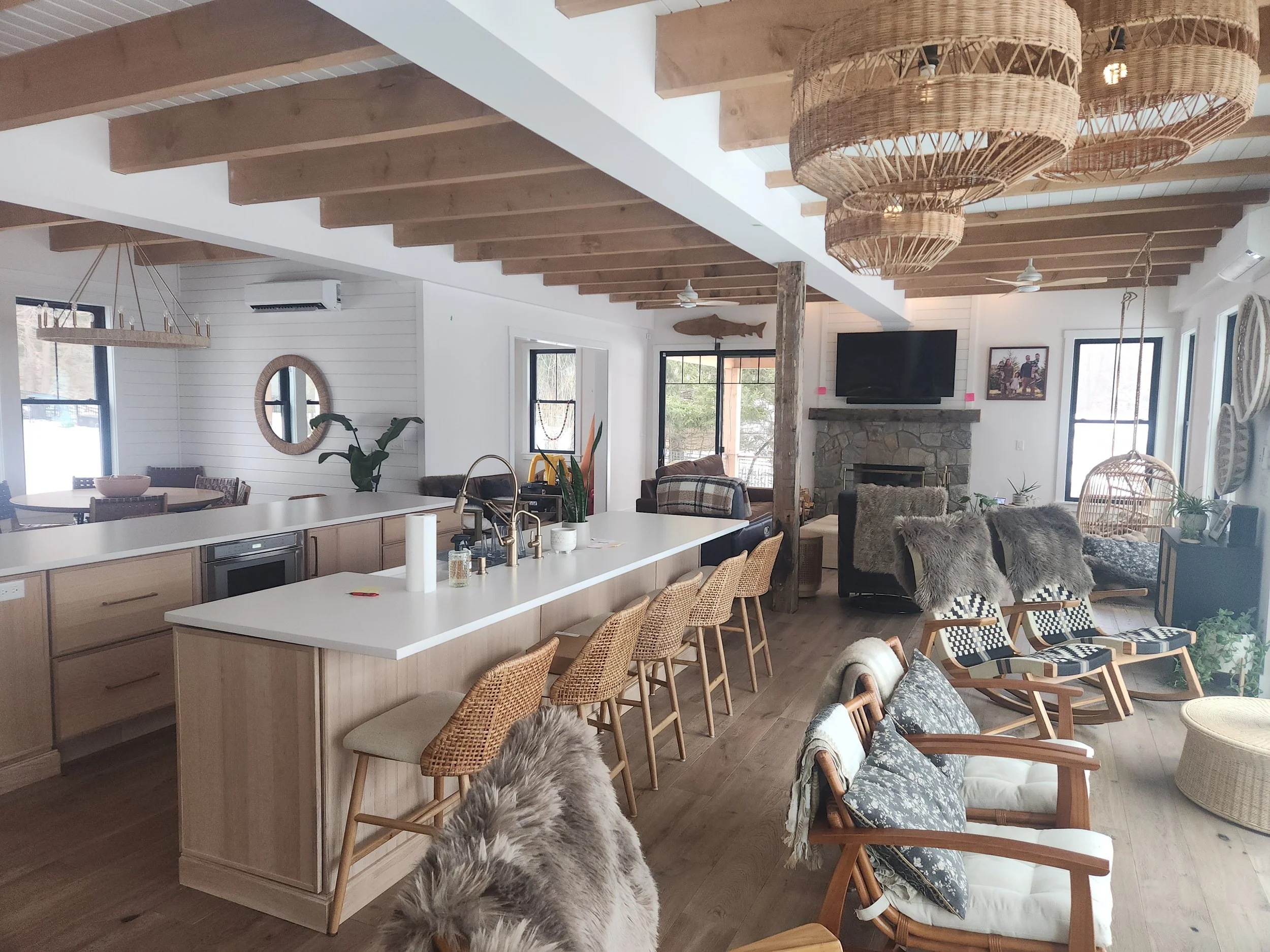 Living room with wooden beams on ceiling, wicker chandeliers, white walls, windows, fireplace, flat-screen TV, various chairs including reading chair with blanket, indoor plants, and a kitchen island with bar stools, and a dog with furry ears in the 