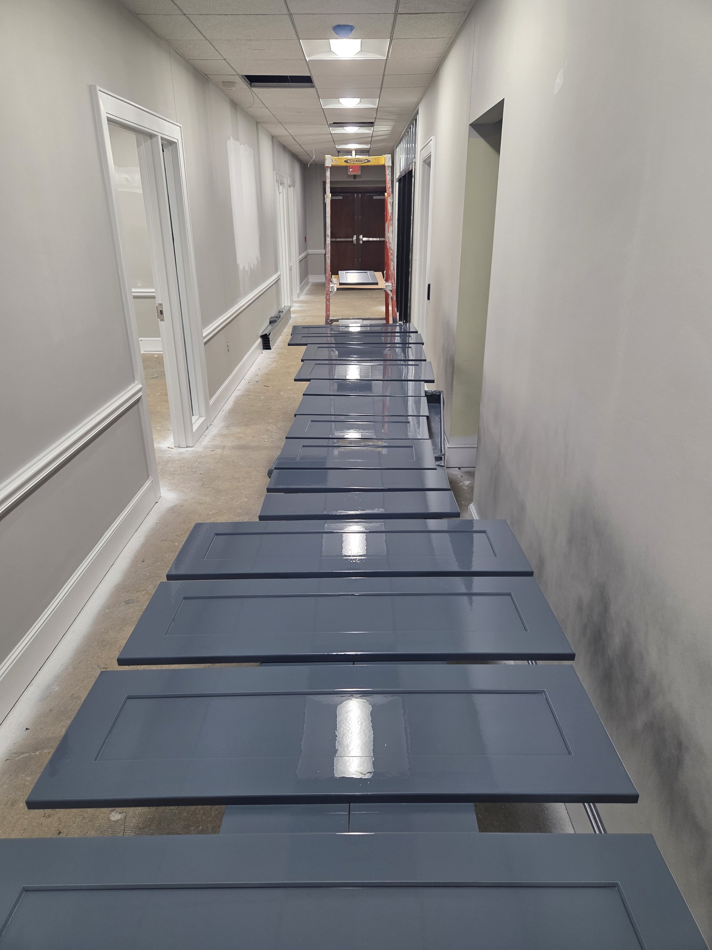 Hallway with gray doors, white walls, ceiling tiles, and a ladder in the center, with gray chairs stacked against the wall on the right.