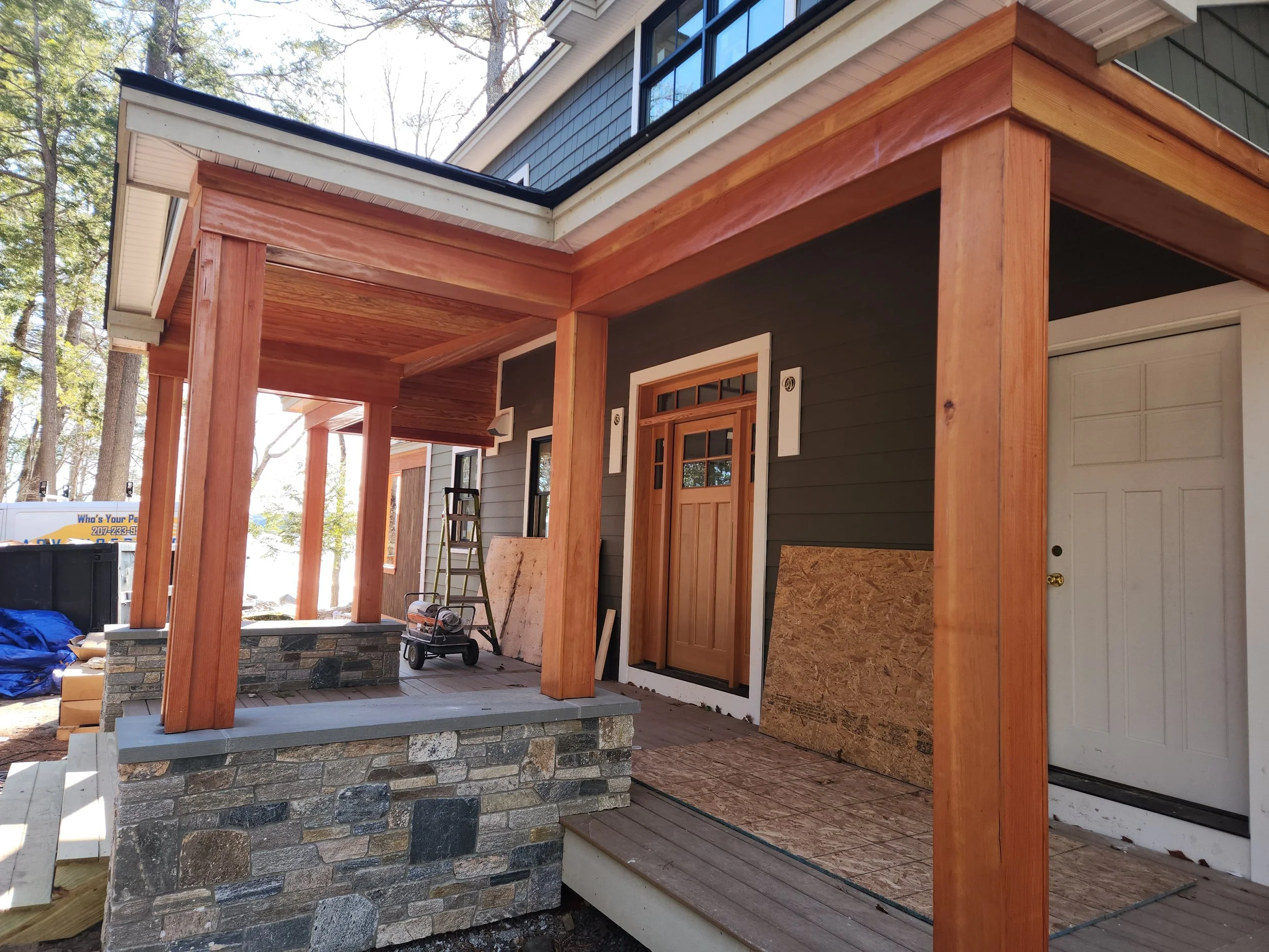 Exterior of a house under construction with a wooden porch, stone foundation, and partially completed siding, surrounded by trees.