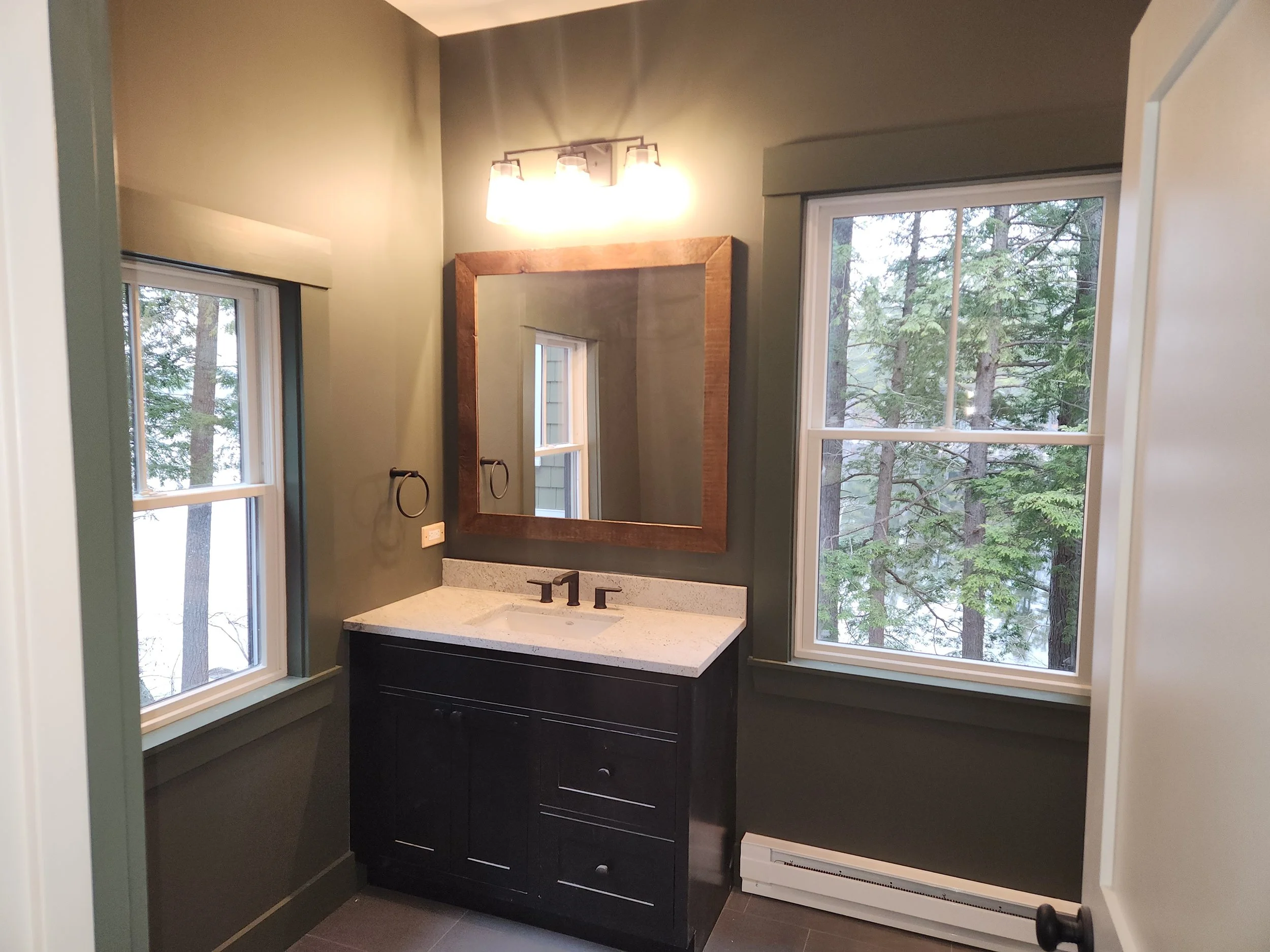A bathroom with large windows showing trees outside, a dark-colored vanity with drawers, a white countertop with a basin sink, a mirror with a wooden frame, and a modern light fixture above the mirror.