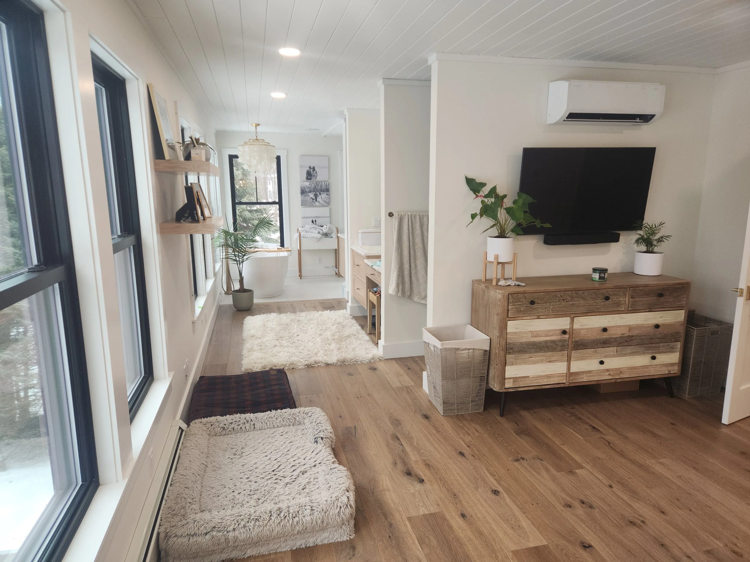 A cozy living space with large windows on the left, two plush white rugs on a wooden floor, and a wooden sideboard with a potted plant and small decorative items. In the background, there is a bathroom with a bathtub, framed artwork, and a hanging li