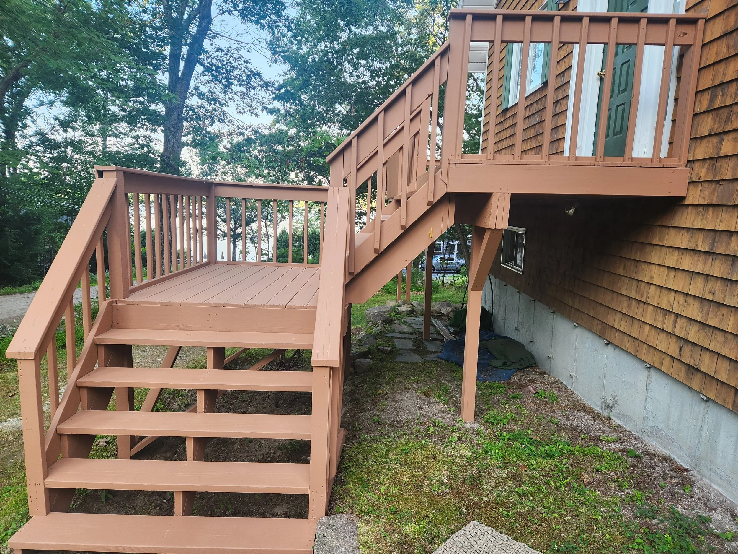 Wooden staircase leading up to a deck beside a house with brown shingles and a green door. Trees with green leaves are in the background.