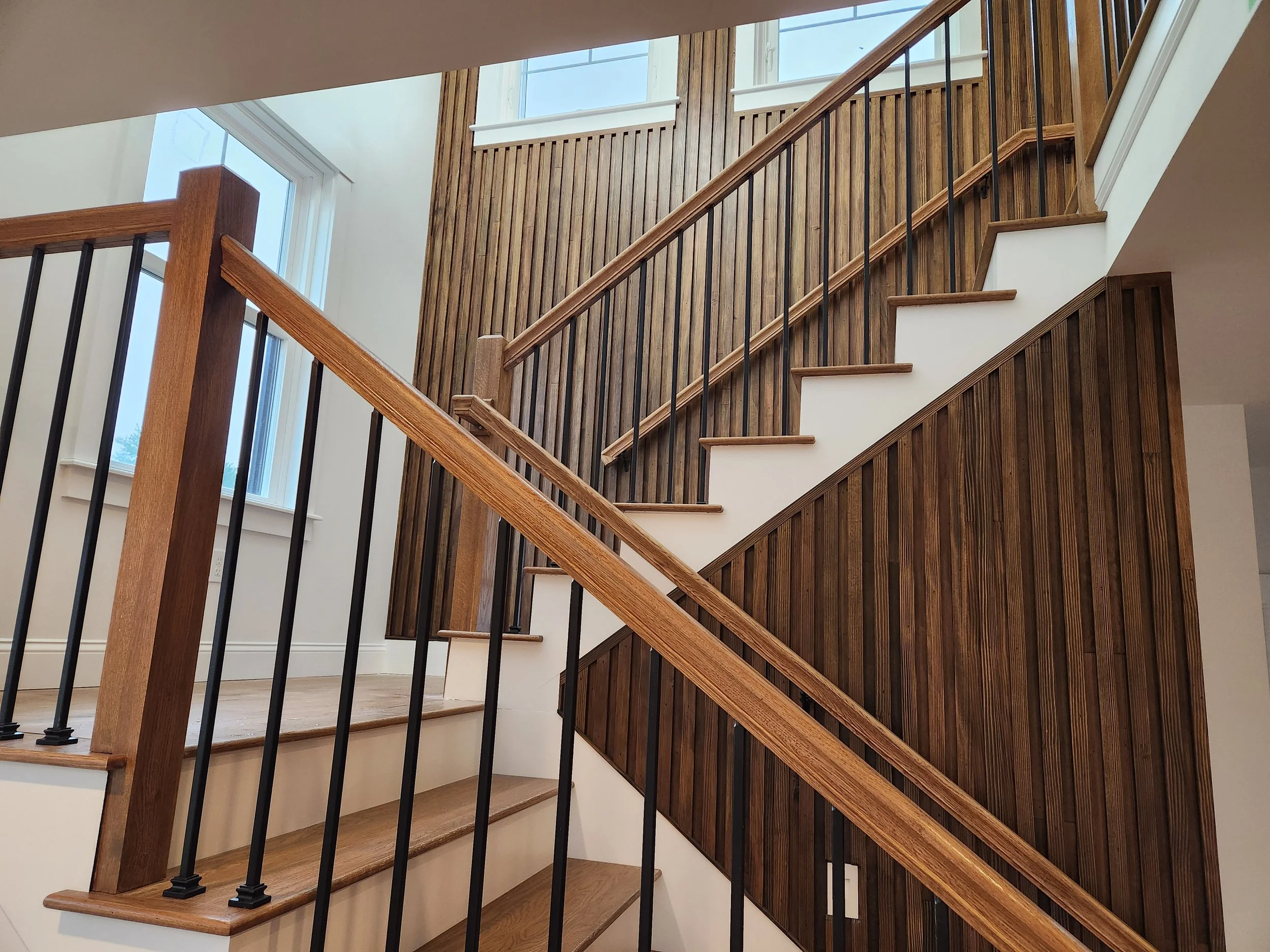 Interior view of a wooden staircase with a wood-paneled wall and large windows letting in natural light.
