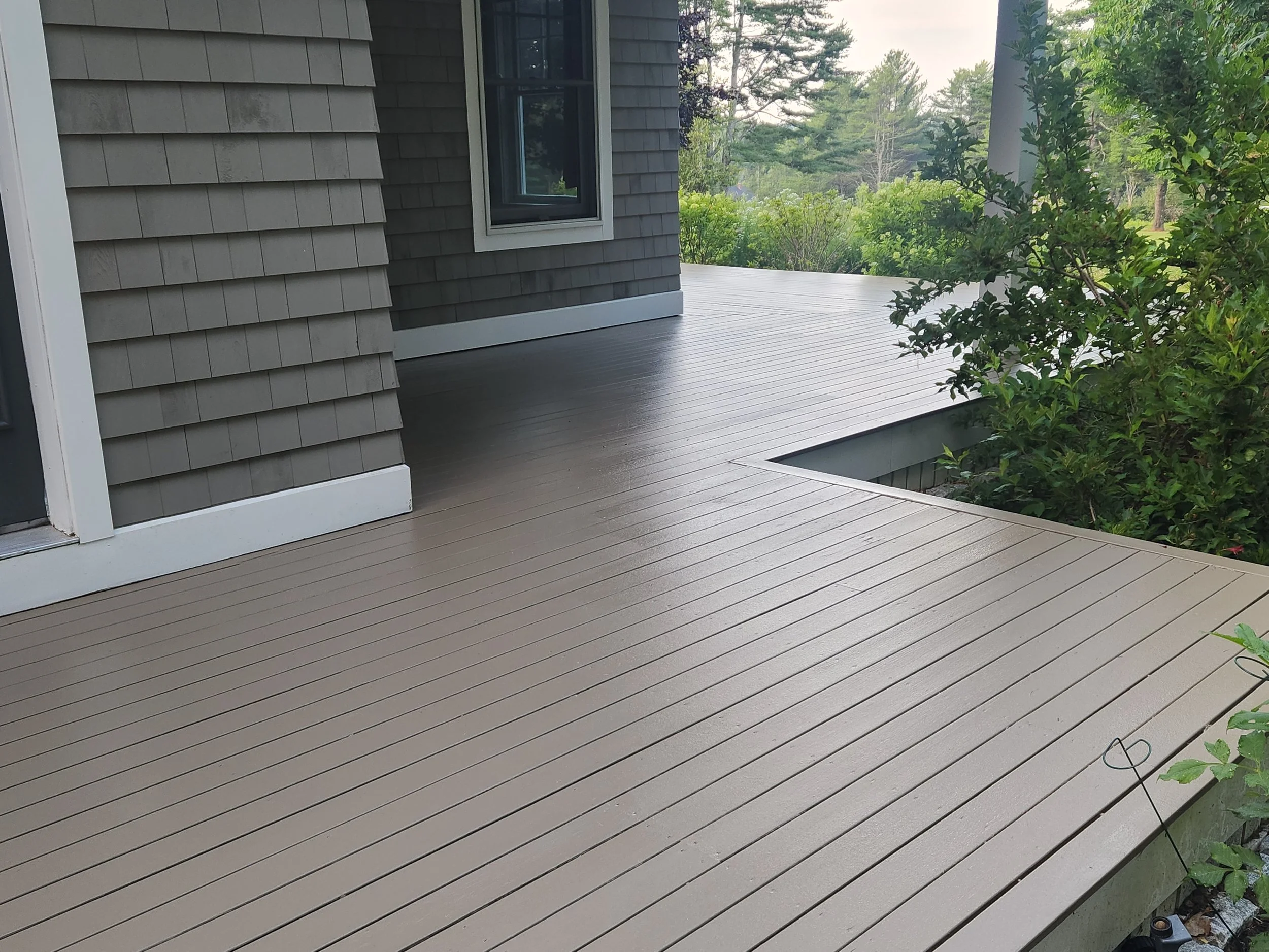 A backyard porch with gray composite decking, a house with gray shingle siding, a window, and surrounding greenery including trees and bushes.