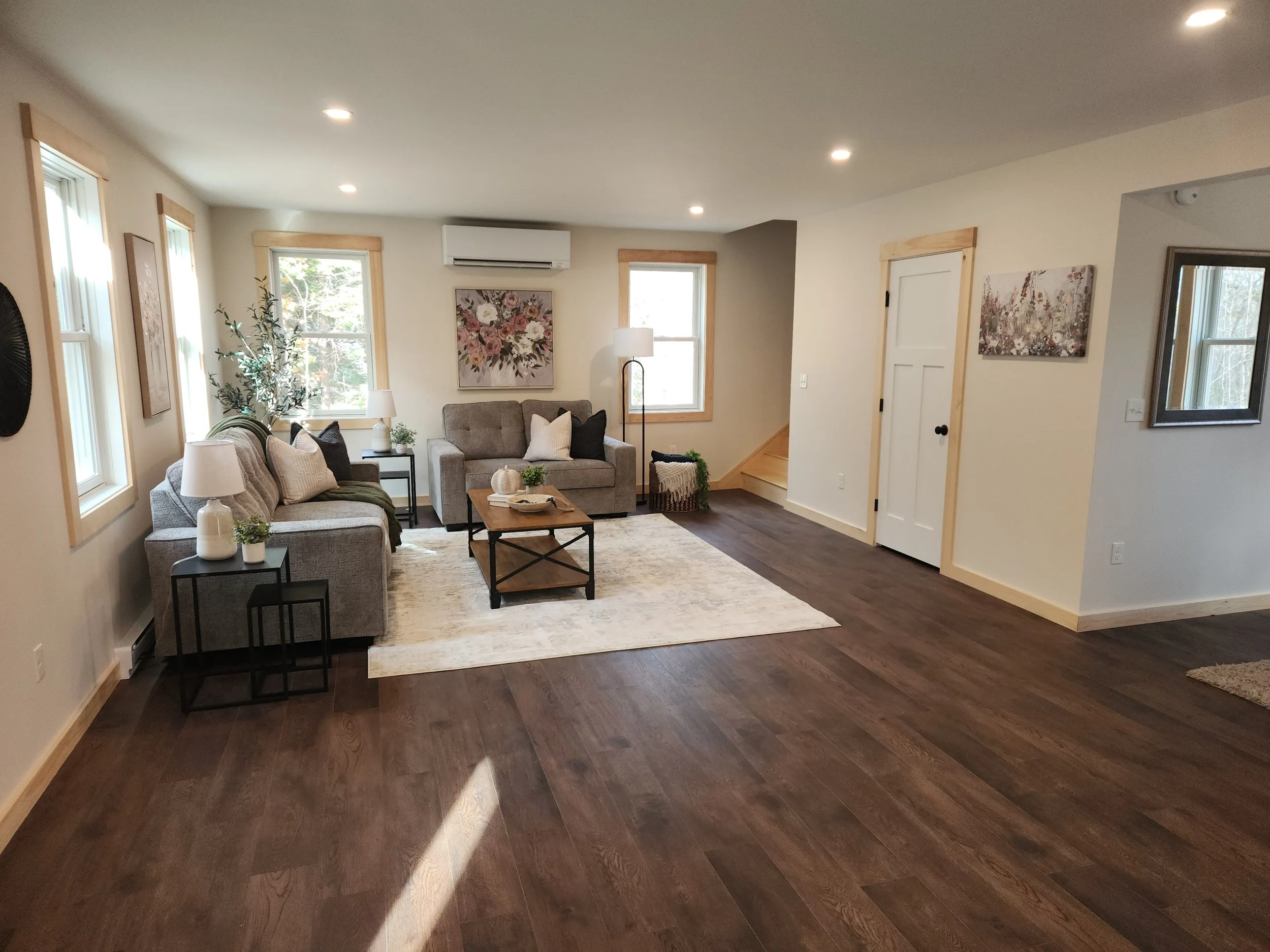 Interior of a living room with hardwood floors, beige walls, multiple windows, and some wall art. There are sofas, side tables, lamps, and decorative plants.