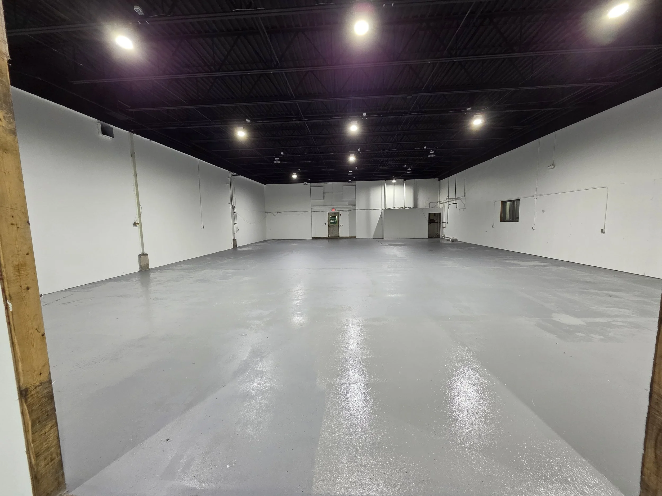 Empty indoor space with polished concrete floor, white walls, black ceiling with fluorescent lights, and visible electrical outlets and piping.