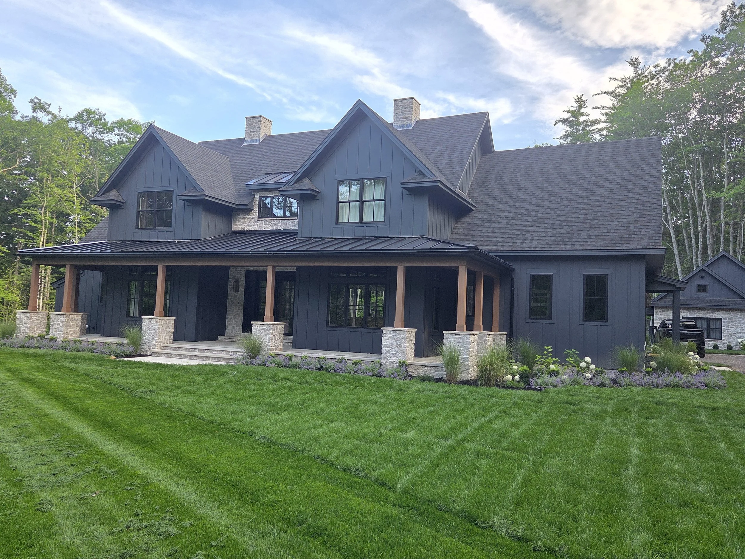 A modern, two-story house painted dark gray with a pitched roof, large windows, a covered porch, and stone columns, surrounded by a well-maintained garden and green lawn with trees in the background under a partly cloudy sky.