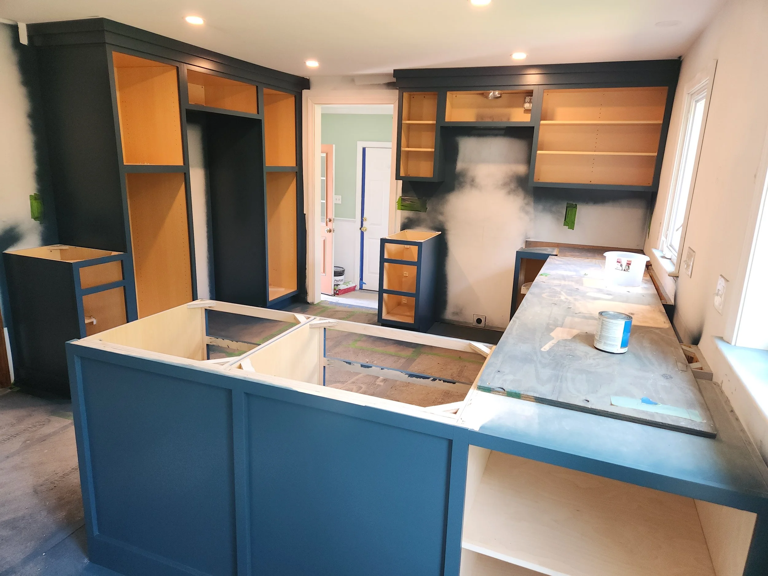 A kitchen under renovation with blue and natural wood cabinets, some opened and some still being installed, a drywall on one wall with painter's tape, a window, and a marble countertop.