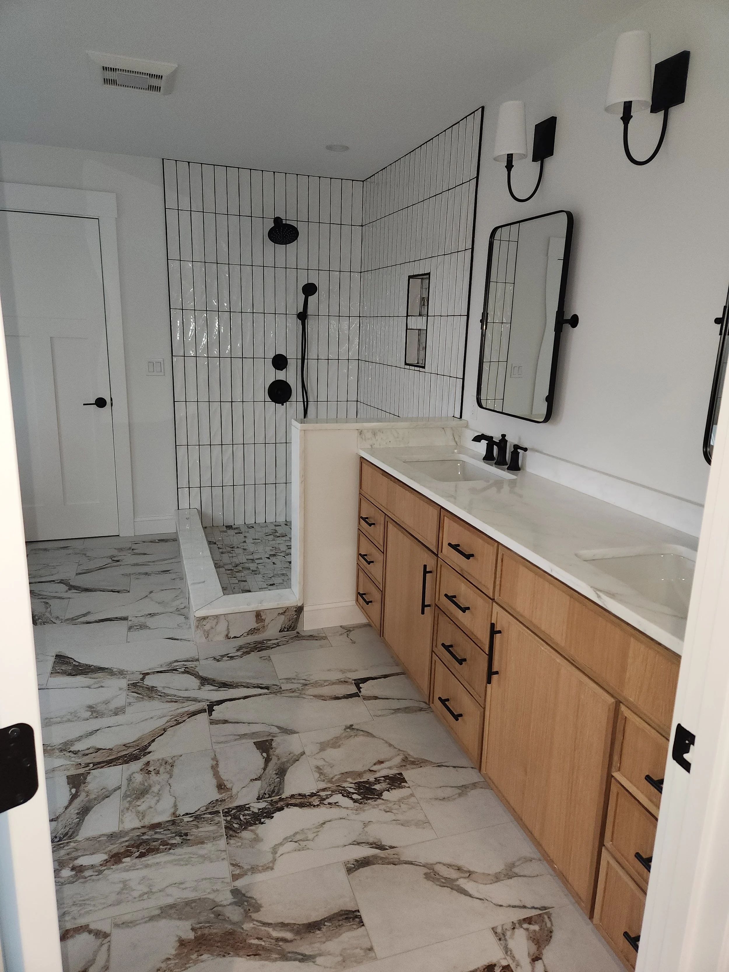 Bathroom with a marble floor, wooden vanity with black hardware, mirror, black wall sconces, and a walk-in shower with black fixtures and white tile walls.