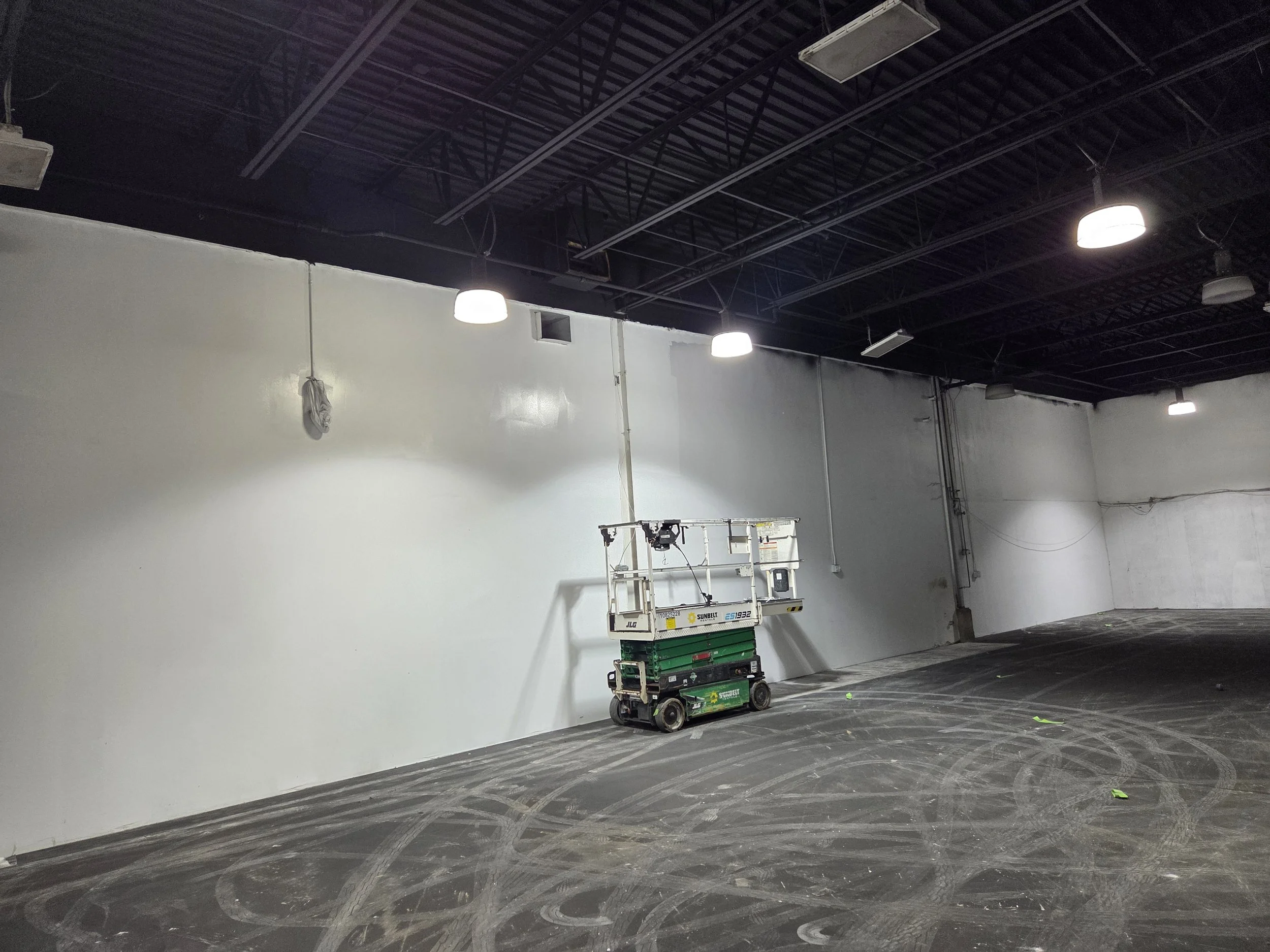 Empty commercial or industrial space with a dark ceiling, bright ceiling lights, white walls, and a concrete floor with tire markings. There is a scissor lift and some green carts on the floor.