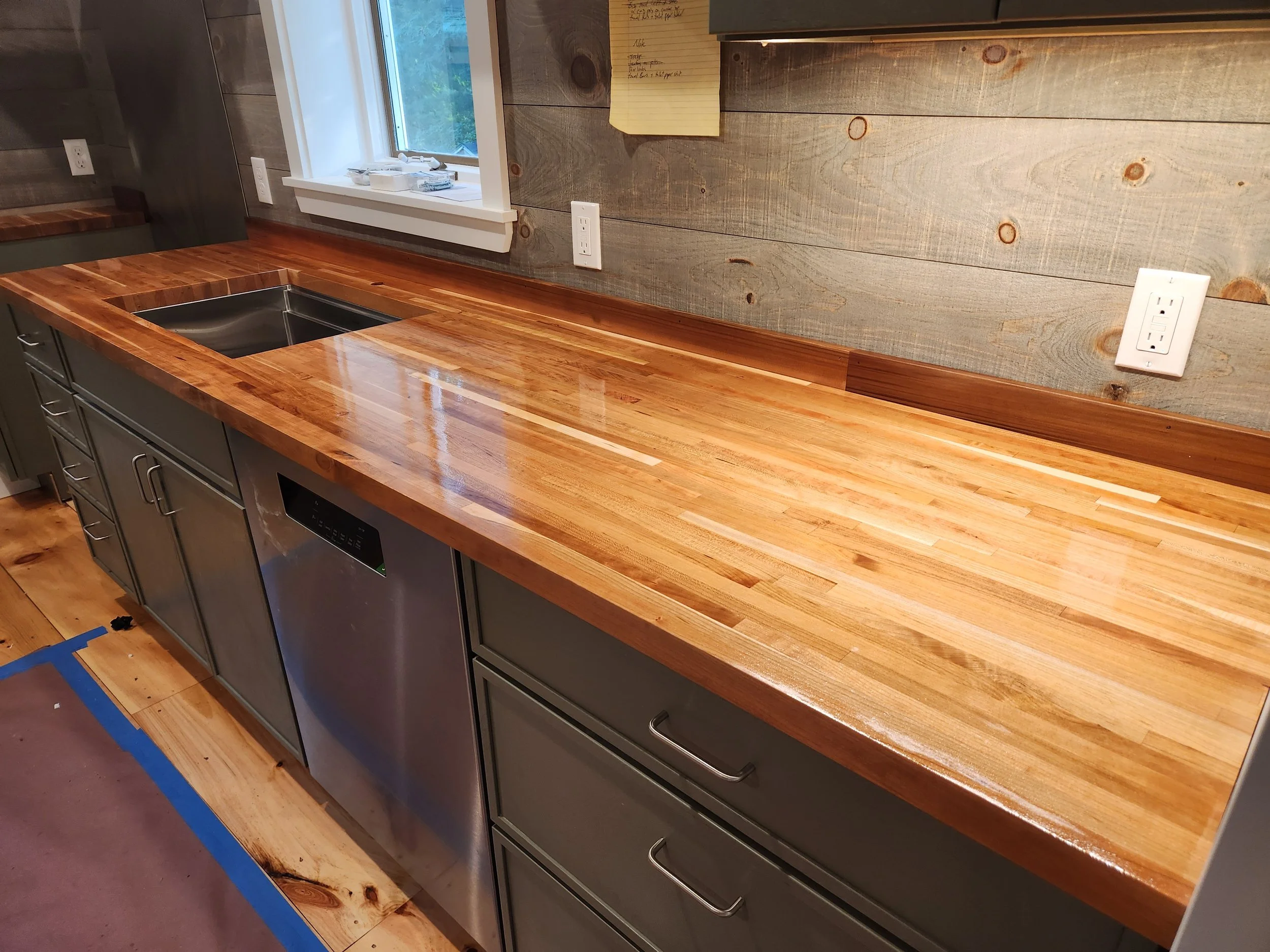 Kitchen with a wooden countertop, gray cabinets, a window above the counter, and electrical outlets on a wooden wall. There is a piece of yellow notepad on the wall.