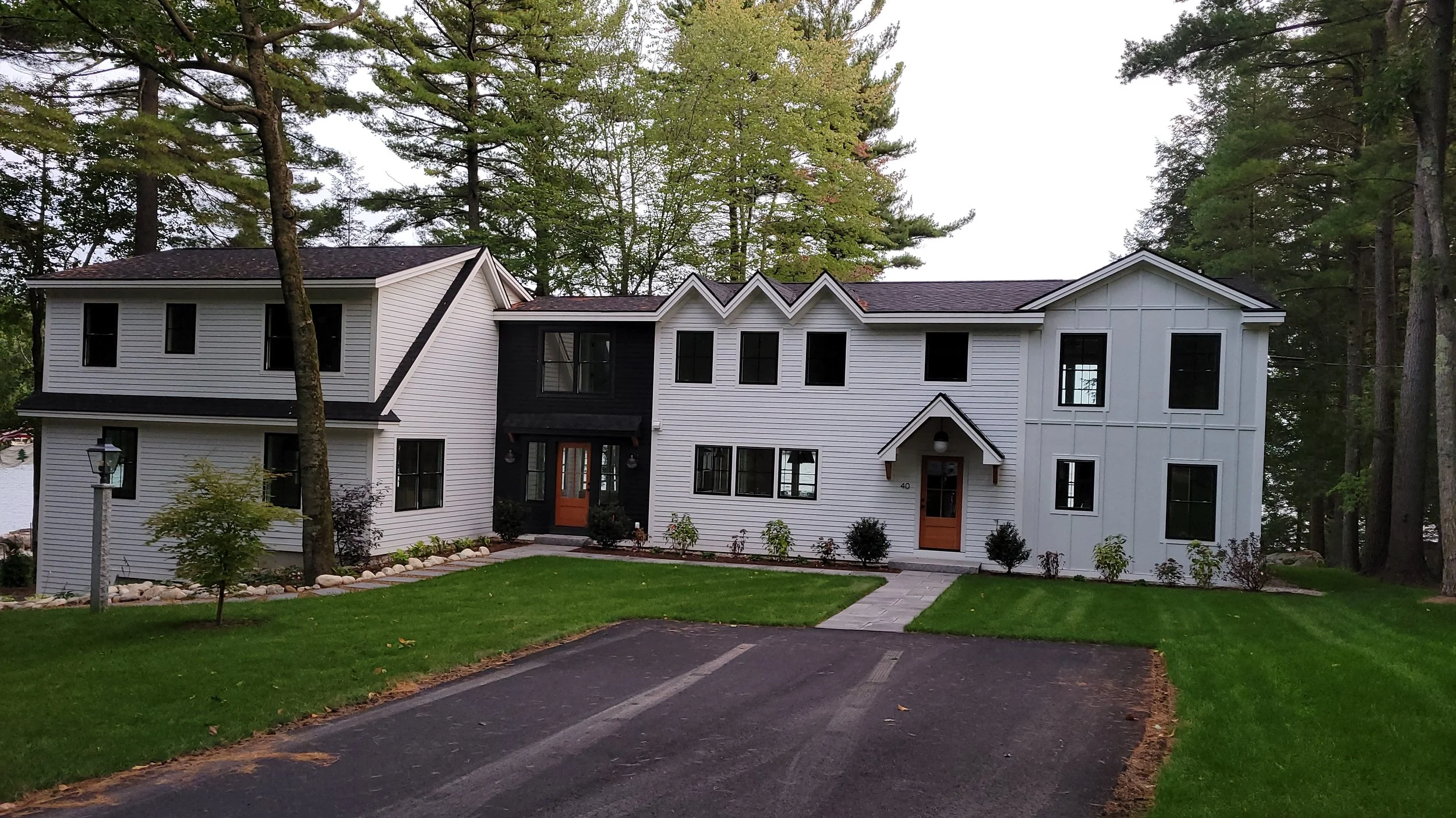 A modern white multi-story house with black accents, located in a wooded area, with a well-maintained lawn, stone walkway, and a parking area in front.