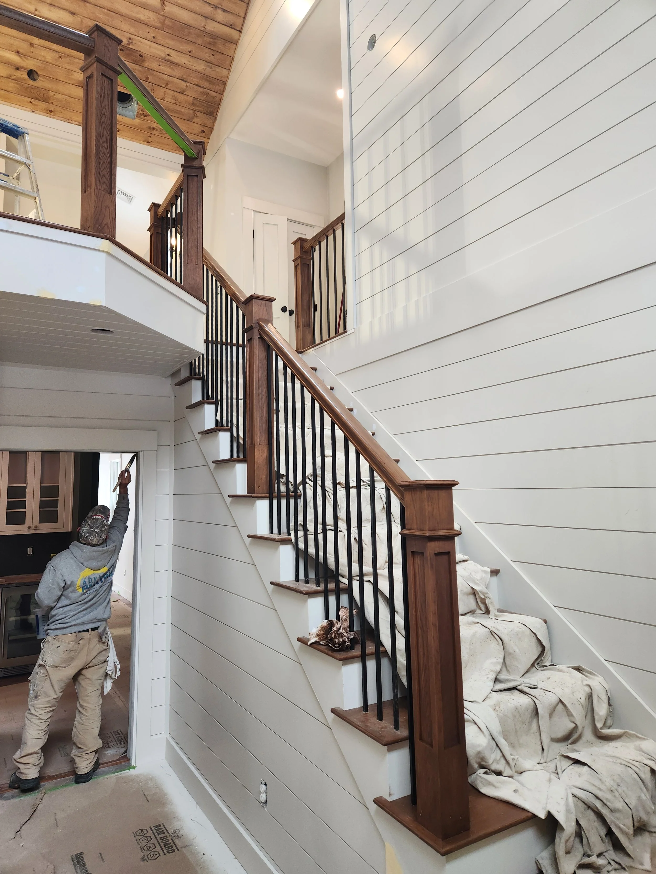 Interior of a house under construction showing a staircase with wooden handrails and black metal balusters, unfinished walls with white paneling, and workers finishing the paint and woodwork.