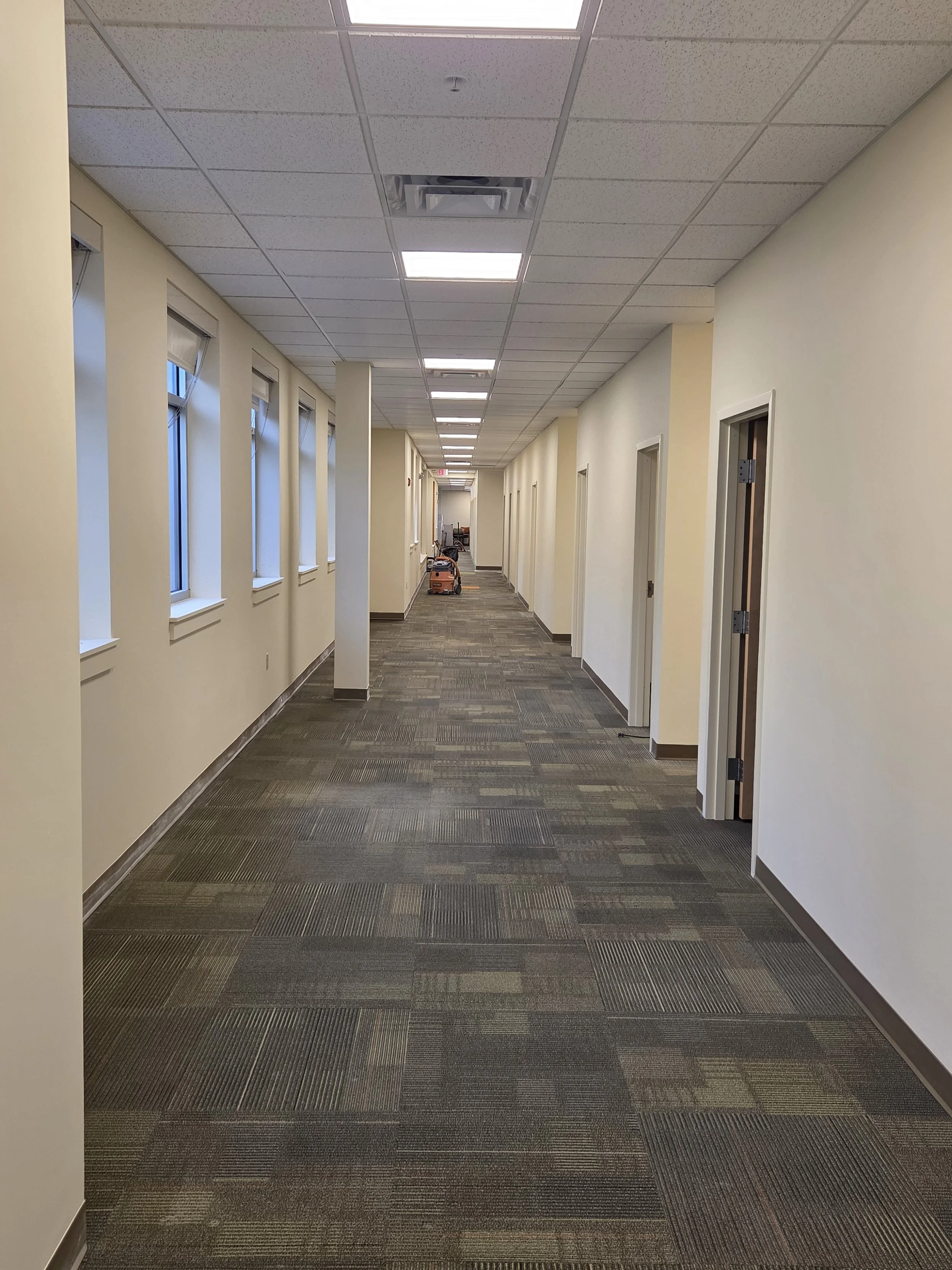 Empty office hallway with windows on the left, white walls, and a brown patterned carpet. Fluorescent lights on the ceiling and an orange cleaning machine in the distance.
