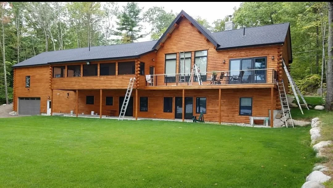 Large wooden house with a black roof, multiple windows, and a balcony with outdoor furniture, surrounded by a grassy lawn and trees.