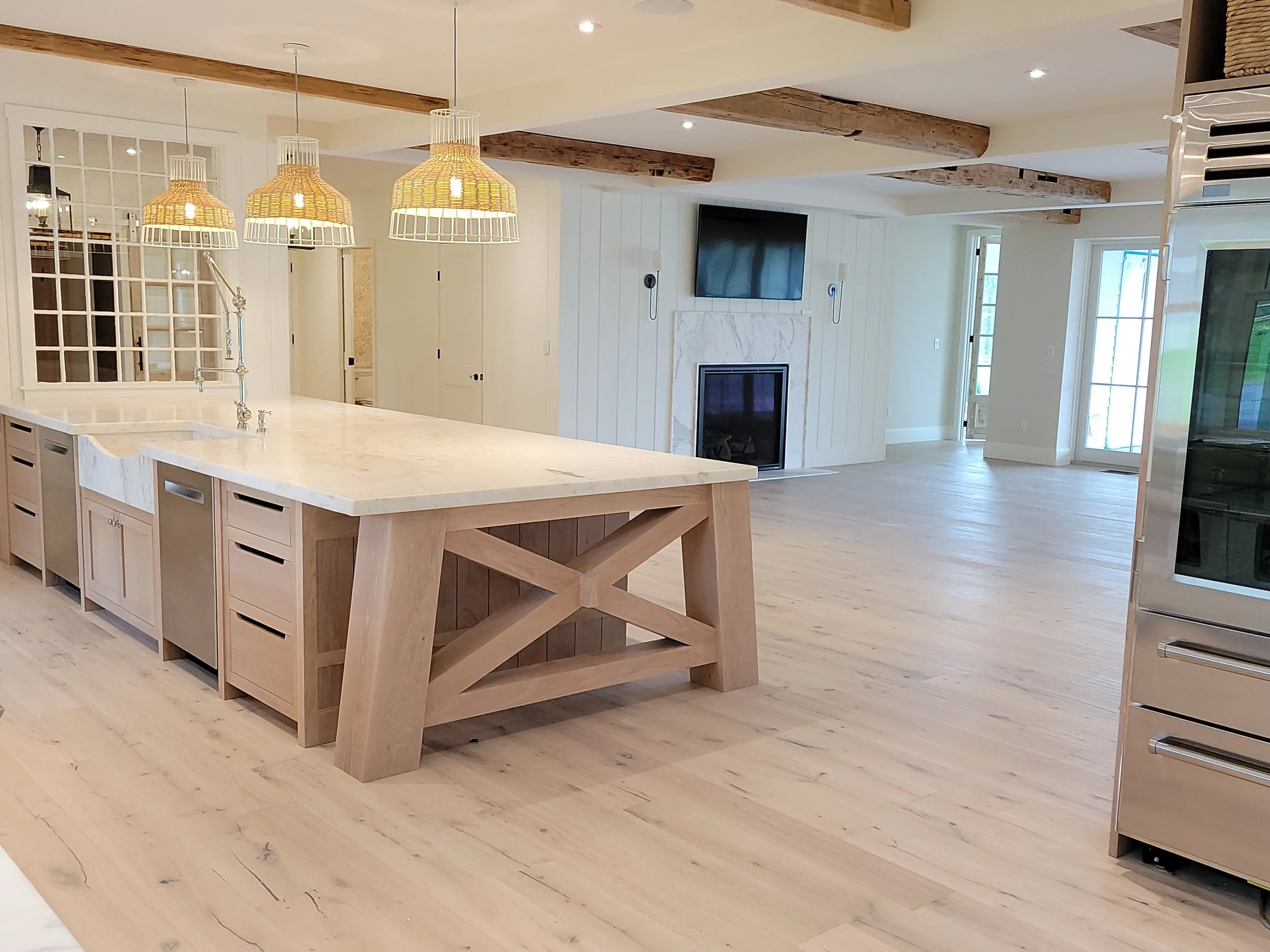 Interior view of a modern kitchen and living area with wooden beams on the ceiling, a white marble kitchen island, pendant lights, a wall-mounted TV, a fireplace, and sliding glass doors.