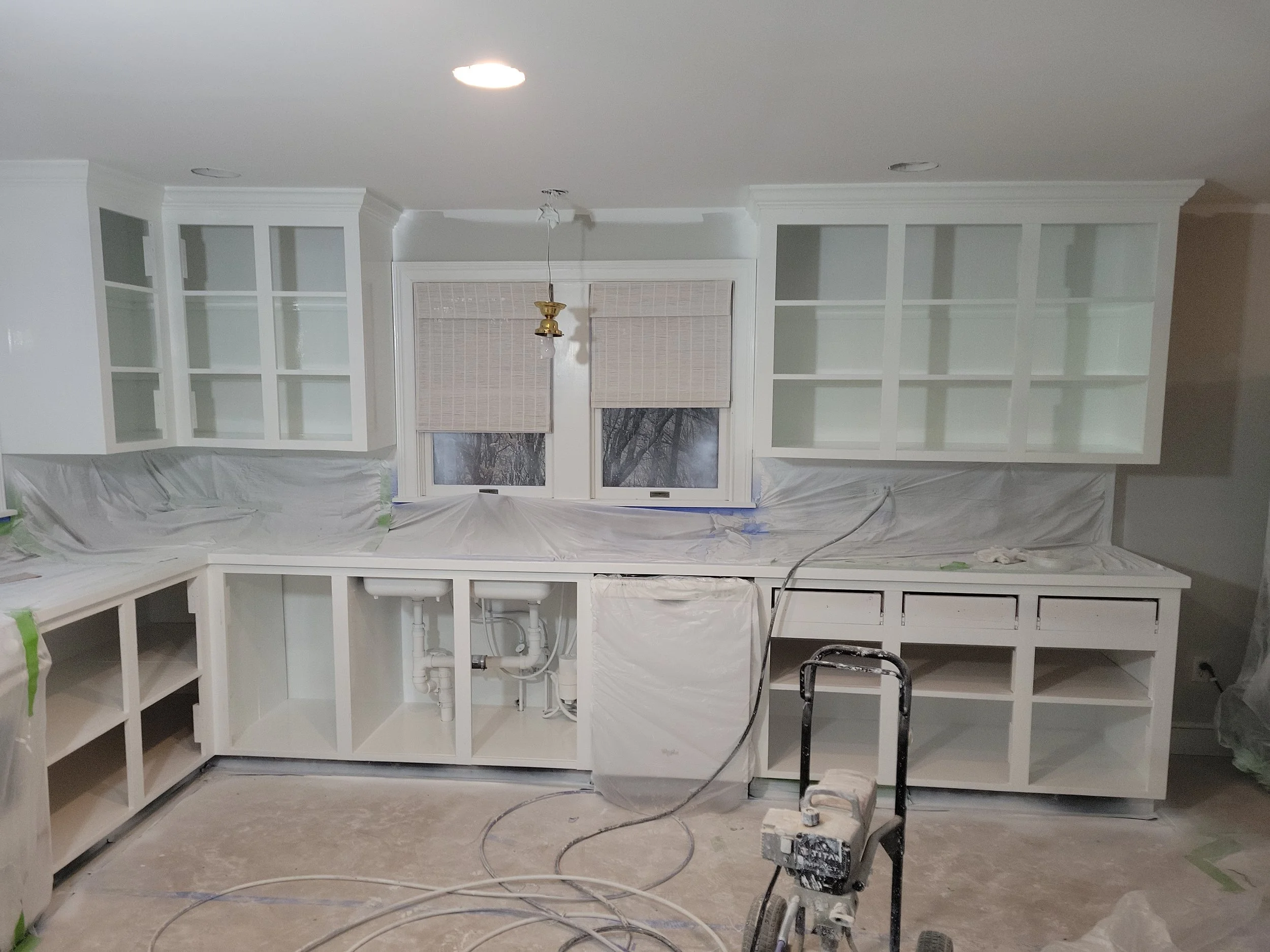 Kitchen cabinets being installed with the lower cabinets unfinished and open, and the upper cabinets painted white. The room appears to be under renovation with tools and protective covering on the floor.