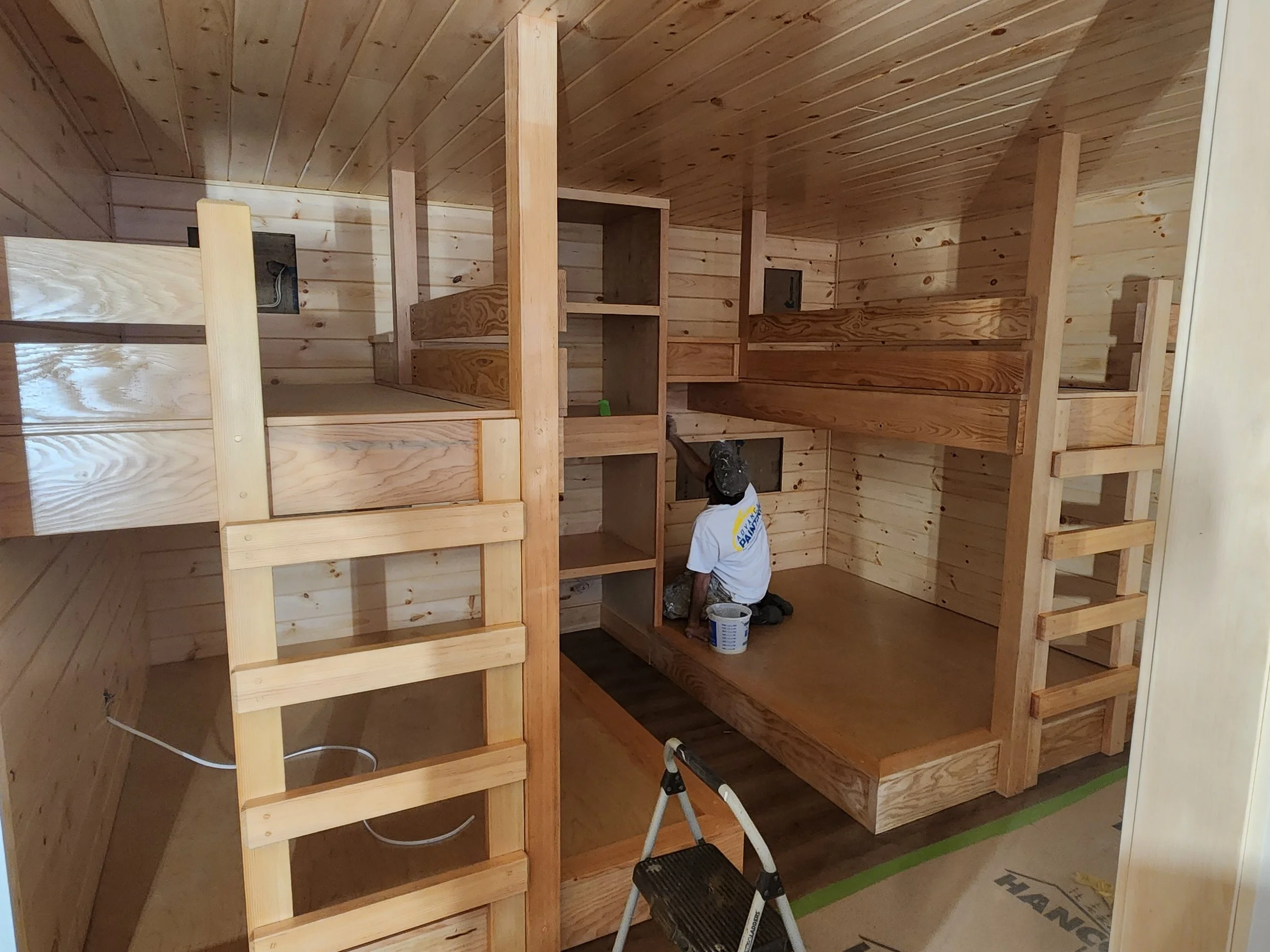 A person working on a carpentry project inside a wooden structure, with light wood framing and paneling, using a step ladder to access higher areas.