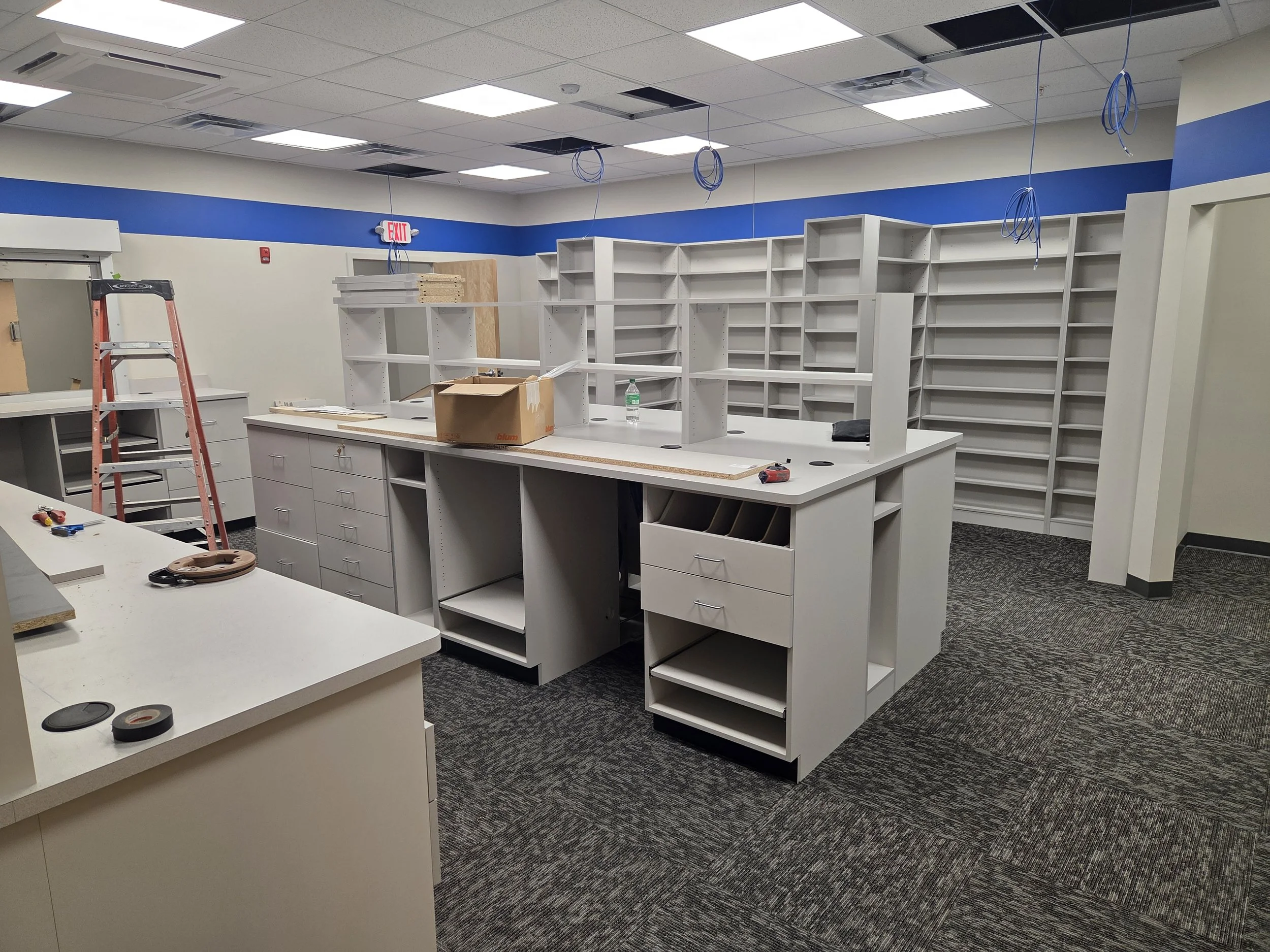An empty office space with white cabinets, shelves, and a reception counter. Some shelves are installed, others are in progress with tools, a ladder, and a water bottle on the counter. Part of the ceiling is missing tiles, and wires hang down.